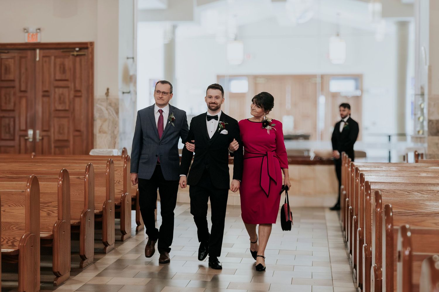 Wedding party members walk down a church aisle between wooden pews.