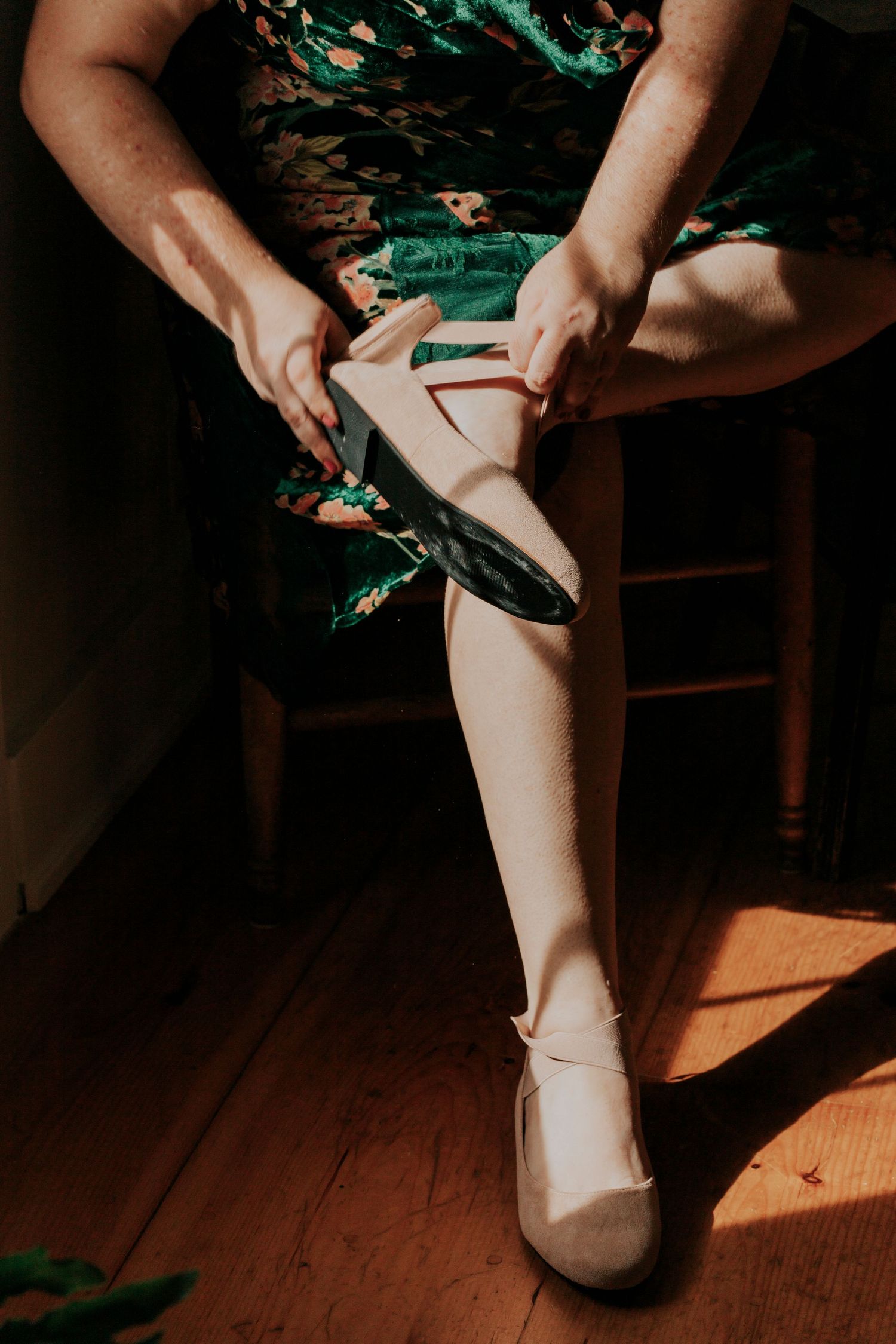 Hands adjusting a pale pink ballet shoe on a foot wearing a floral print dress against a wooden floor in warm lighting.