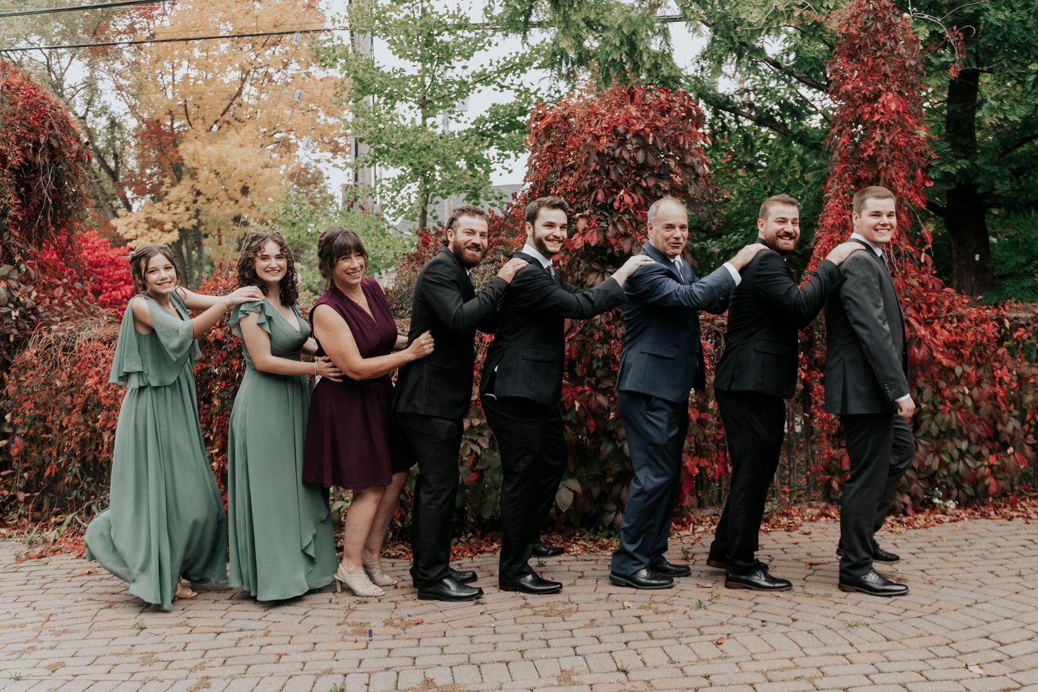 Wedding party poses in a line against autumn foliage wearing sage green dresses and black suits.