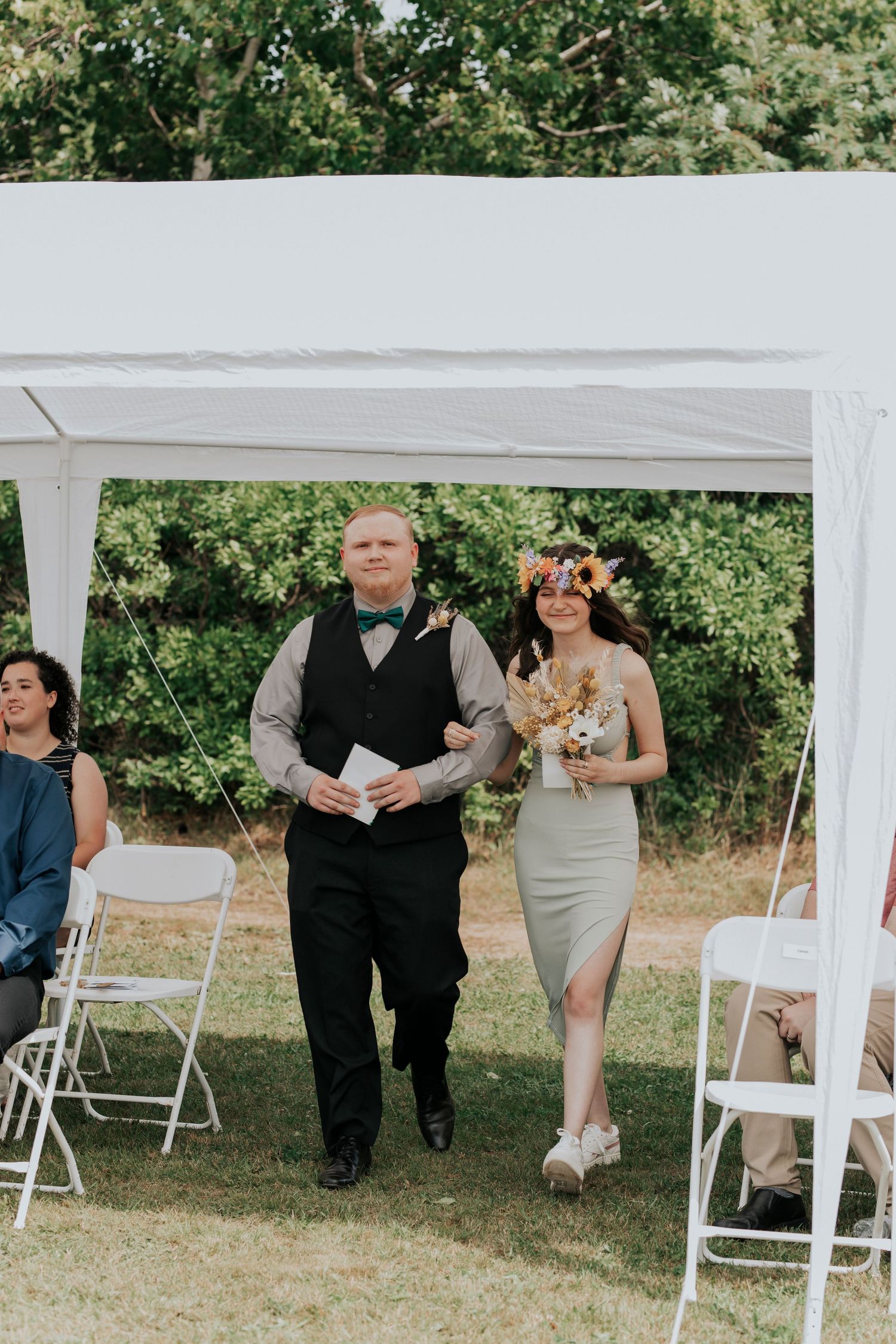 A couple walks under a white wedding tent canopy during an outdoor garden ceremony with folding chairs on the lawn.