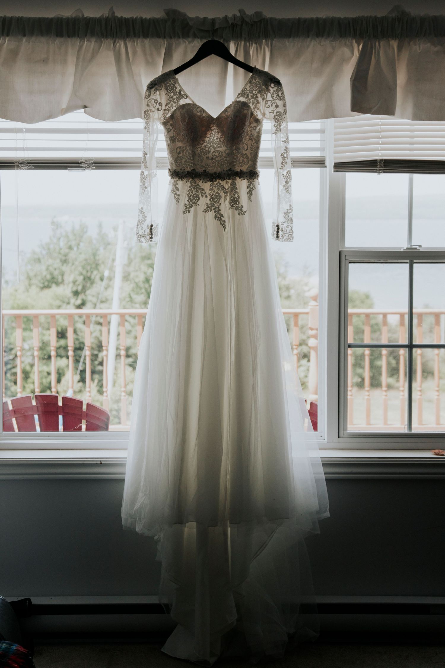 An elegant white wedding dress with lace sleeves and flowing tulle skirt hangs in front of a sunlit window.