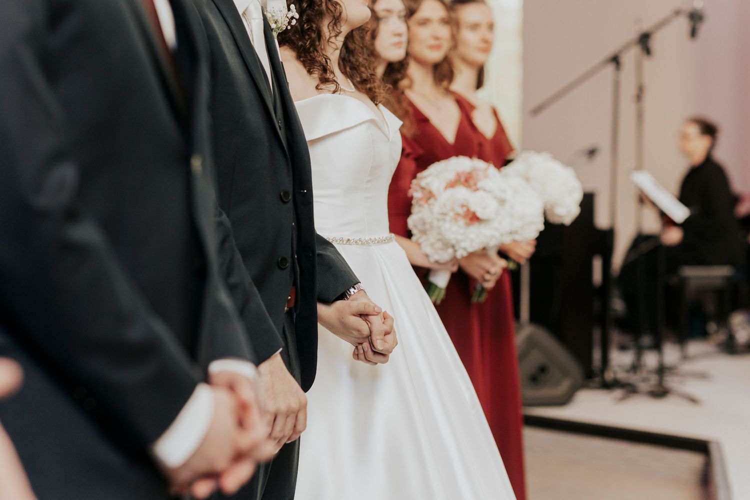 A couple stands at a wedding ceremony with a bridesmaid in burgundy holding a white floral bouquet and piano in background.