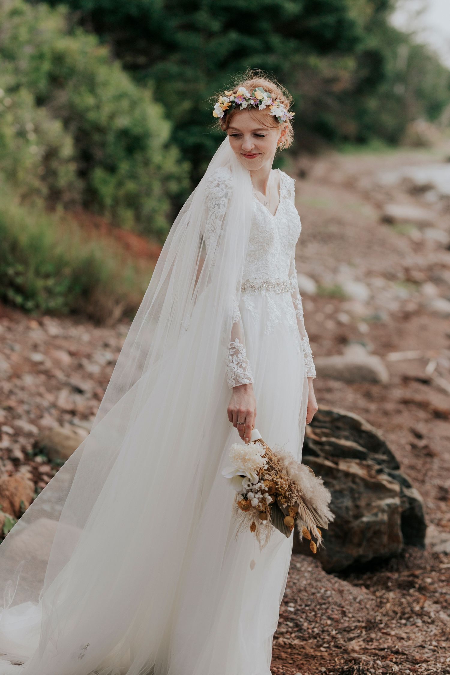 A bride in a vintage-style long sleeve lace wedding dress and floral crown stands on rocky terrain holding dried flowers.