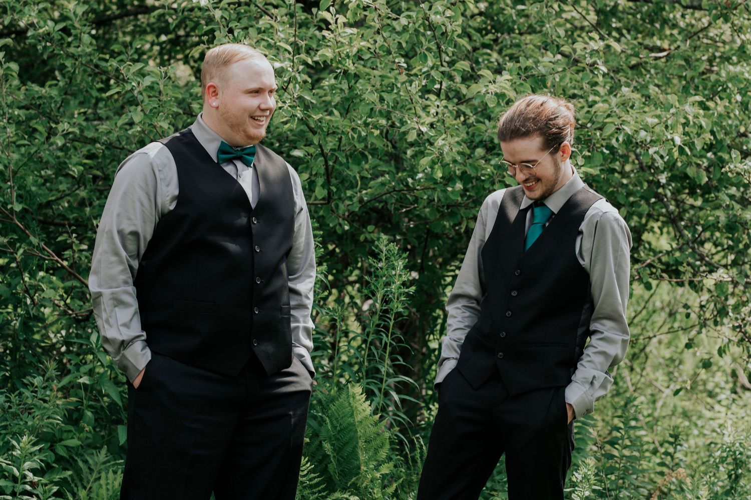 The groom and best man in matching black vests stand together in conversation against a lush green outdoor background.