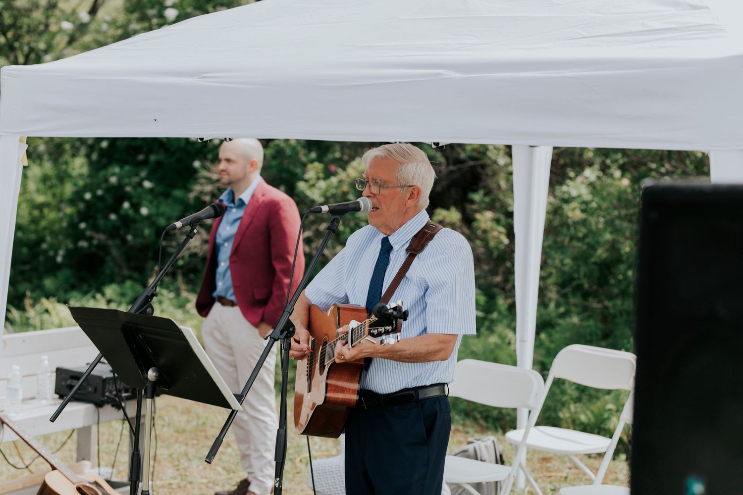 Musicians perform under a white tent canopy at an outdoor event, with one playing guitar and another watching a music stand.