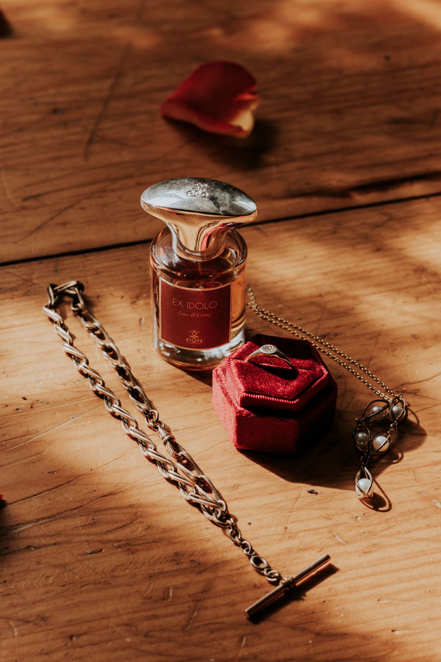 A perfume bottle and red velvet jewelry boxes arranged with a vintage chain bracelet on a wooden floor in warm lighting.