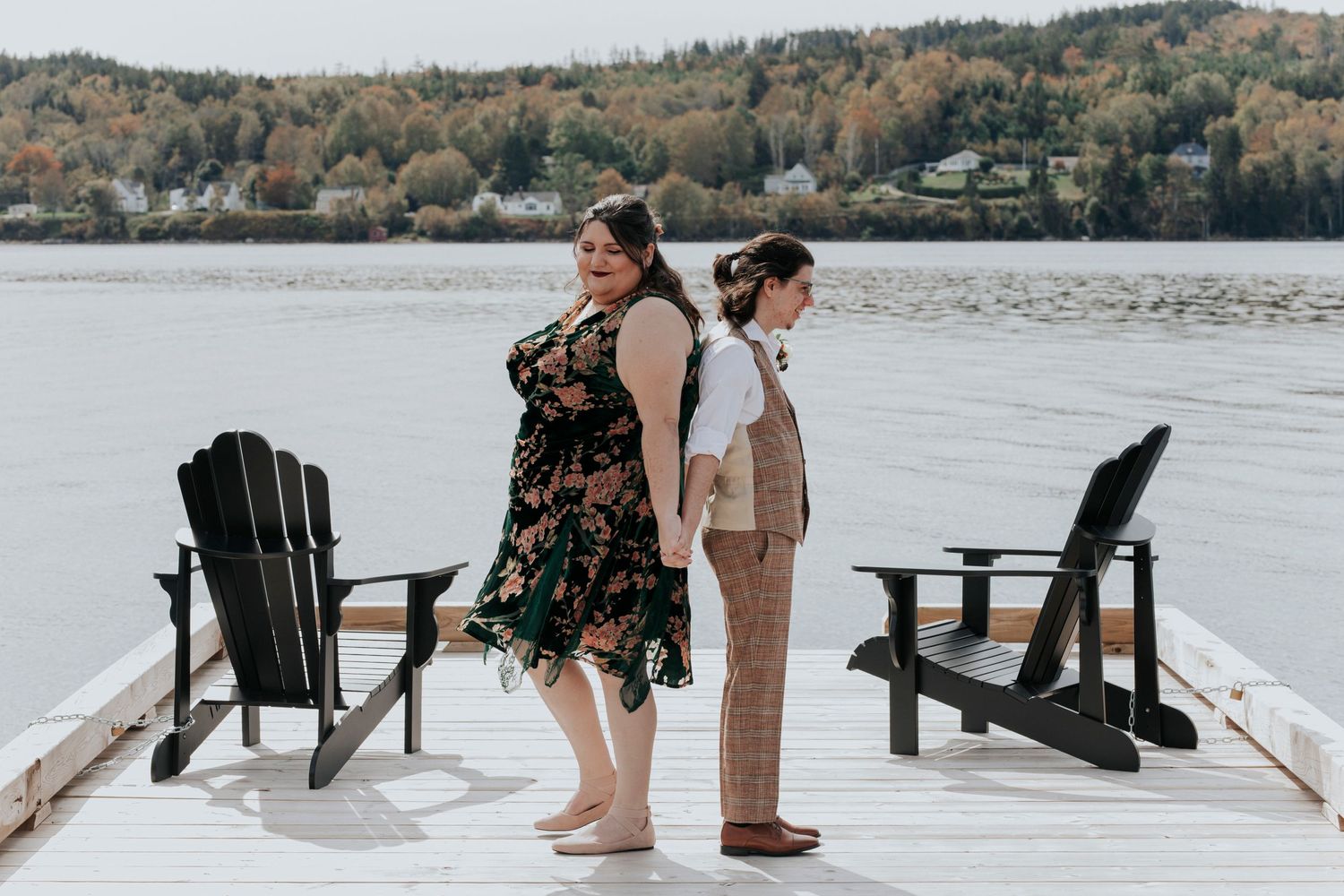 Bride and groom stand back-to-back on a wooden dock with deck chairs overlooking a serene lake and autumn forest before their first look.