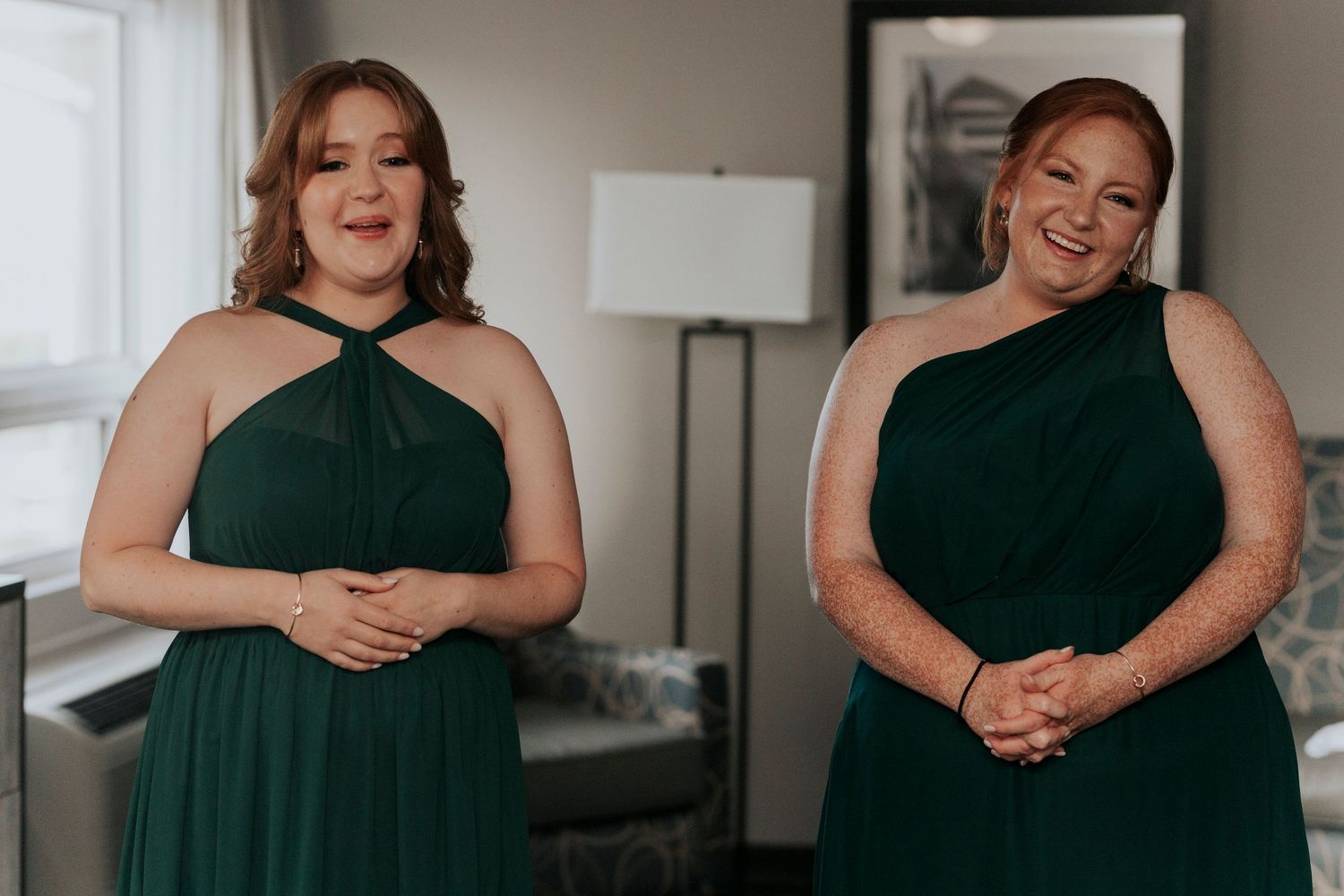Two bridesmaids in elegant emerald green one-shoulder dresses share a joyful moment.