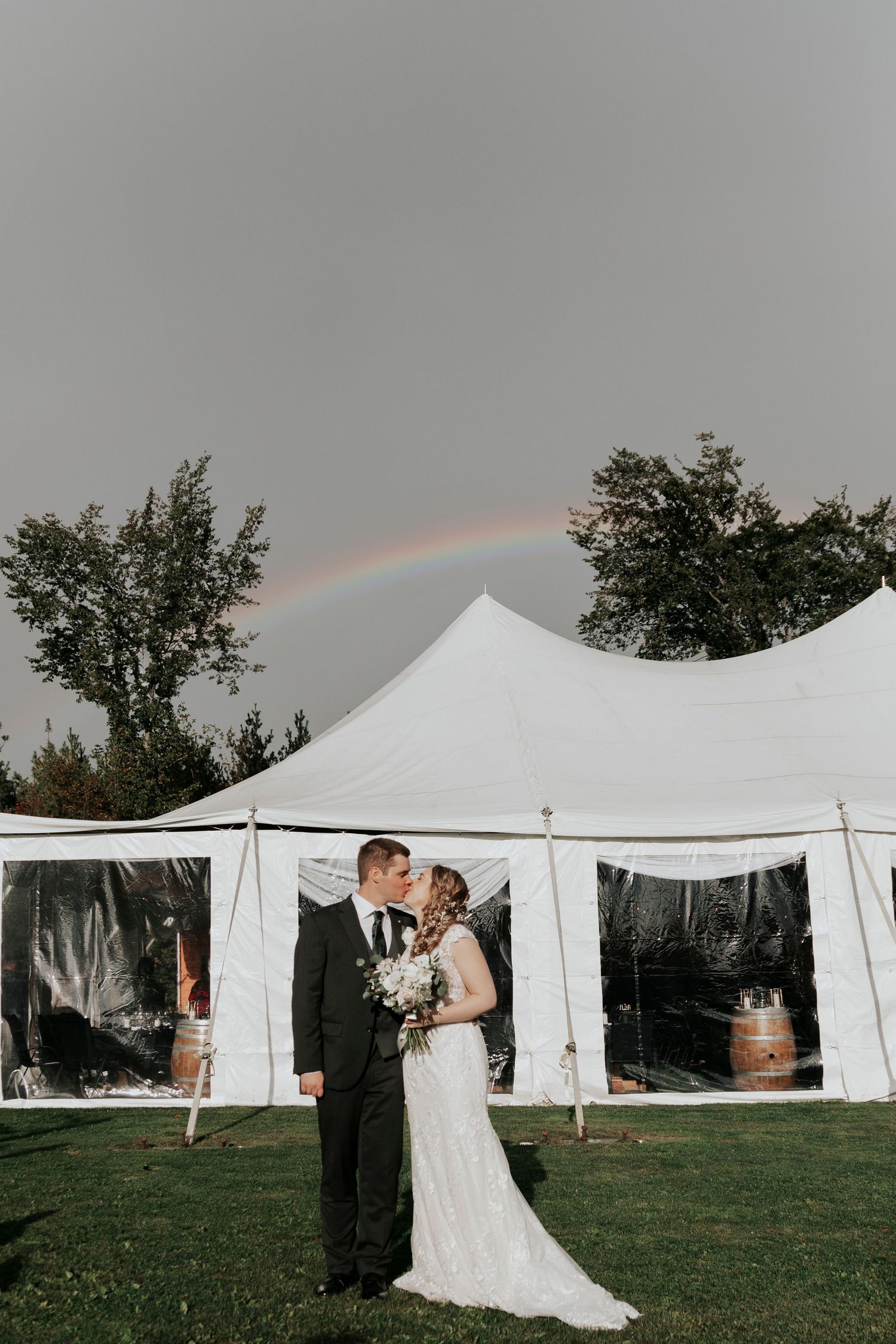 A bride and groom kissing in front of a white wedding tent with a rainbow in the sky.