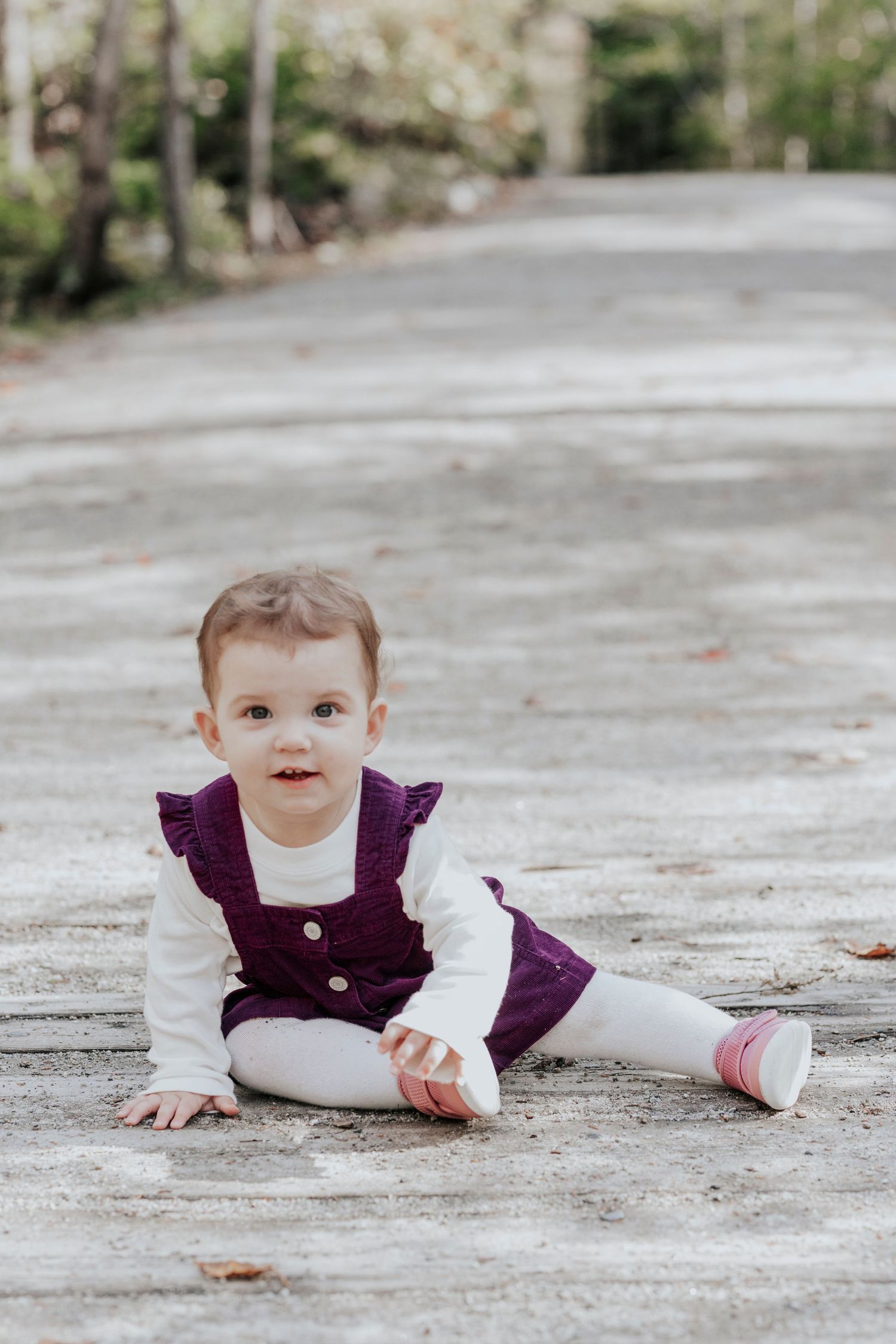 Adorable baby girl in purple dress sitting on a path surrounded by autumn trees.