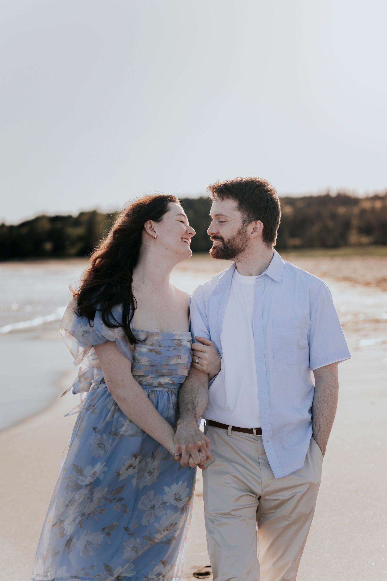 Couple smiling at each other while holding hands and walking along a sandy beach at golden hour.
