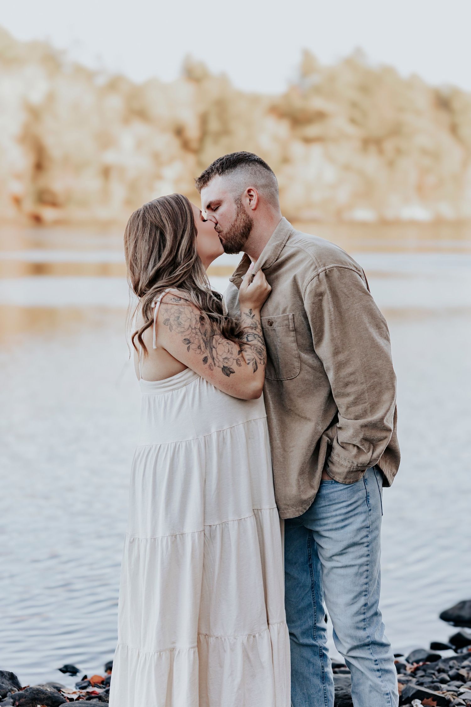 Couple shares a romantic kiss by a serene lake, woman in white maxi dress, man in tan shirt and jeans.