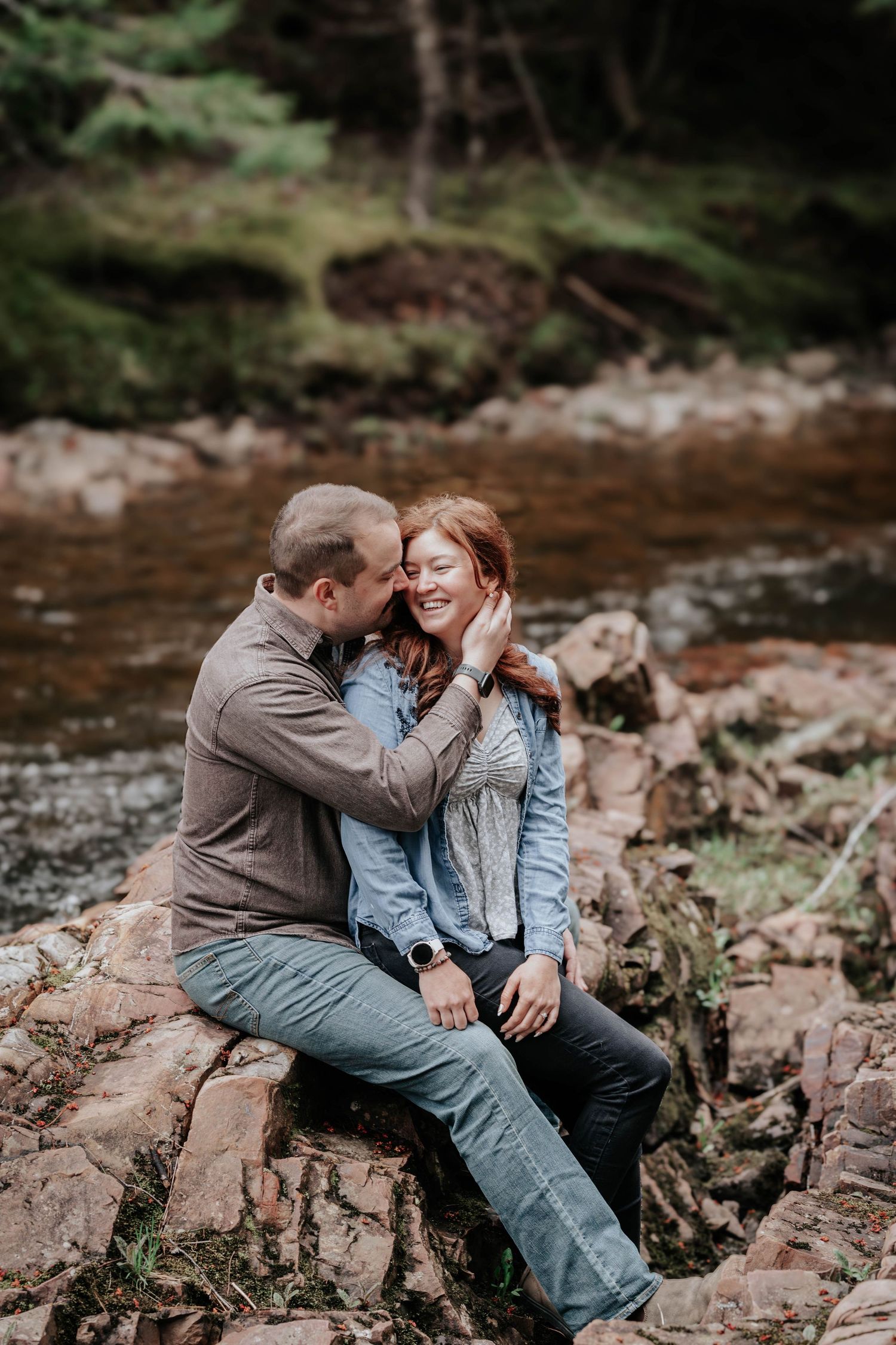 Couple sitting on rocks by forest stream, man gently touching redhead woman's face as she laughs.