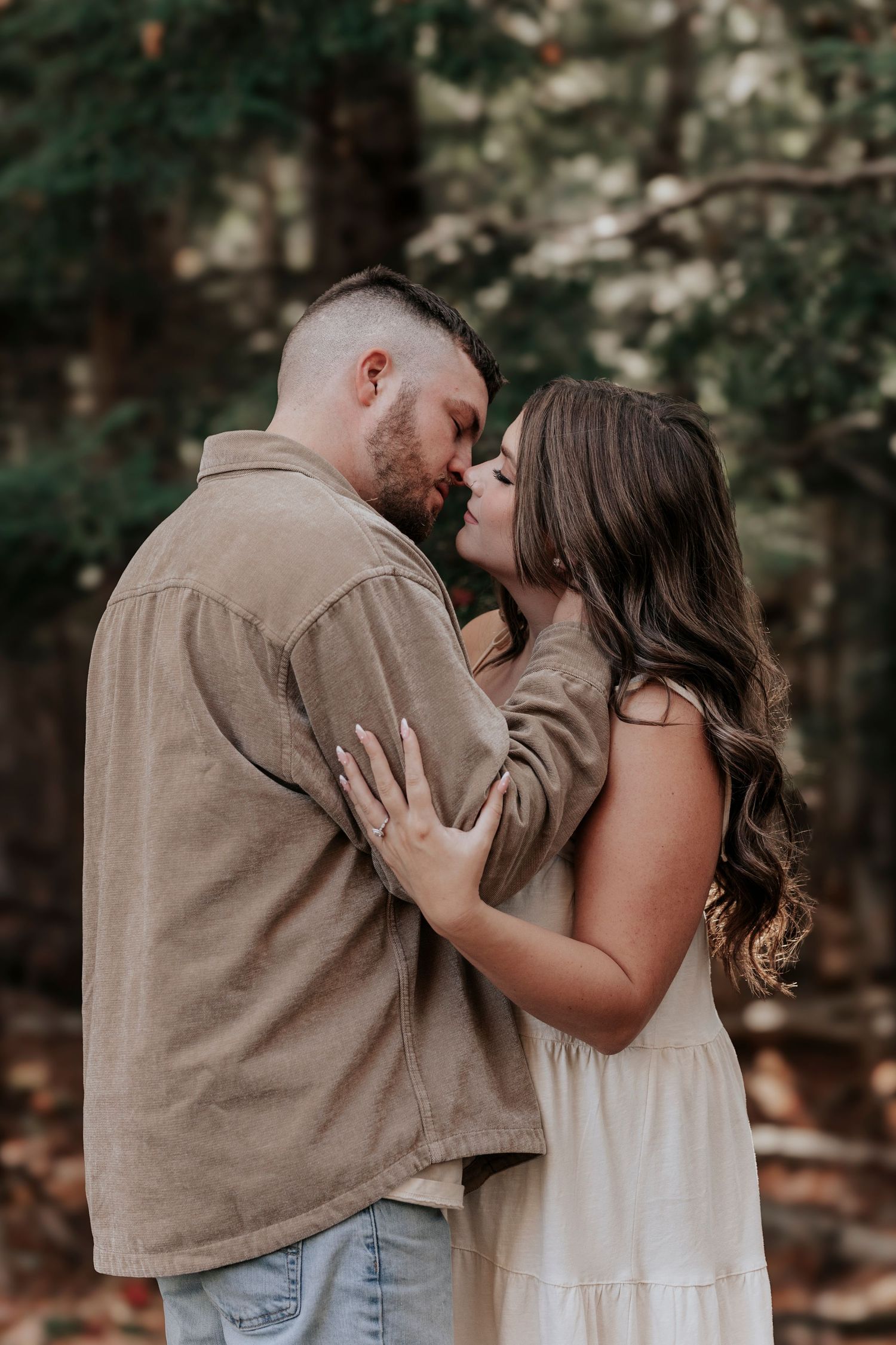 Couple sharing a tender almost-kiss embrace in a forest, man in tan shirt and woman in white dress.