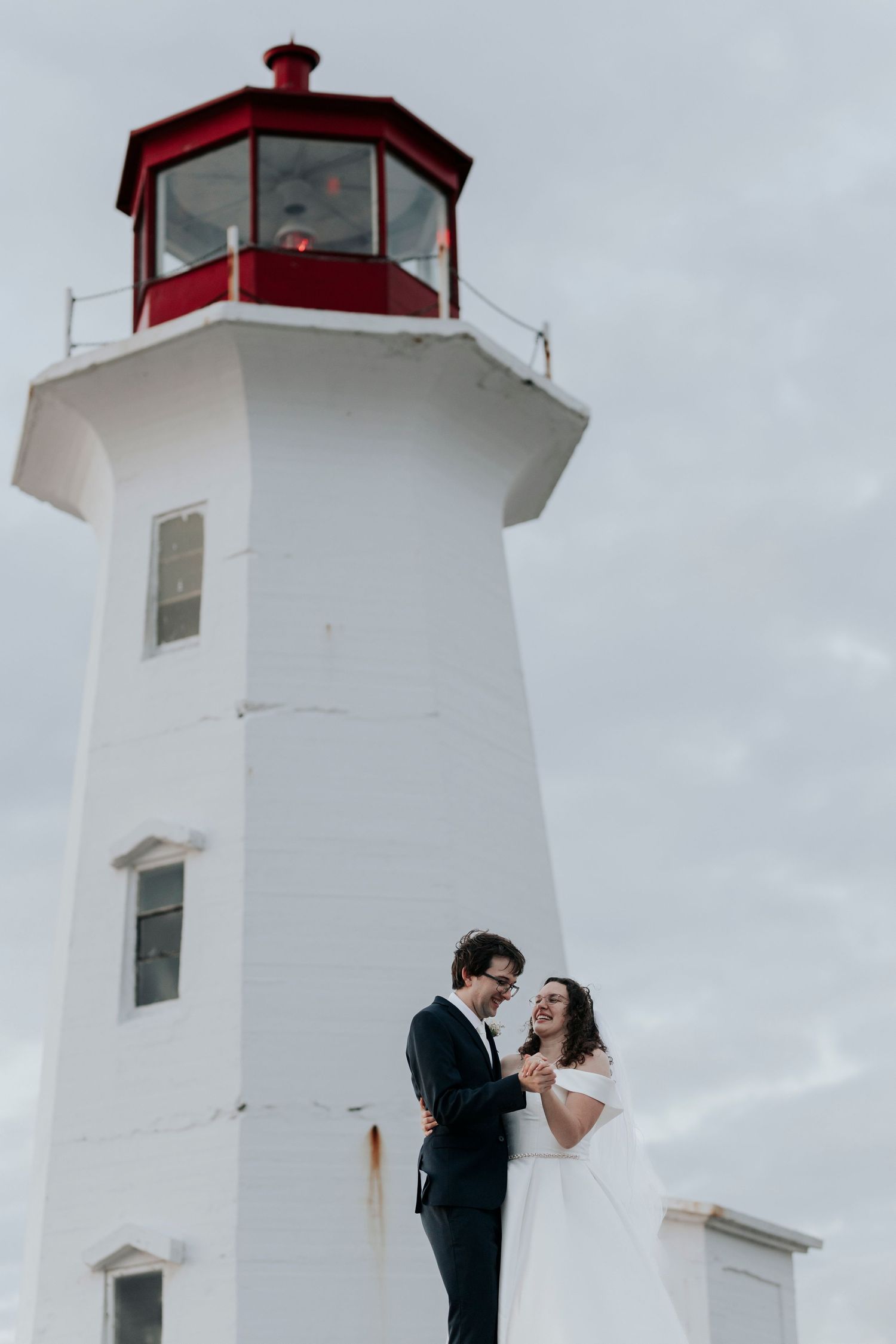Bride and groom share romantic moment beside white and red lighthouse under cloudy sky.