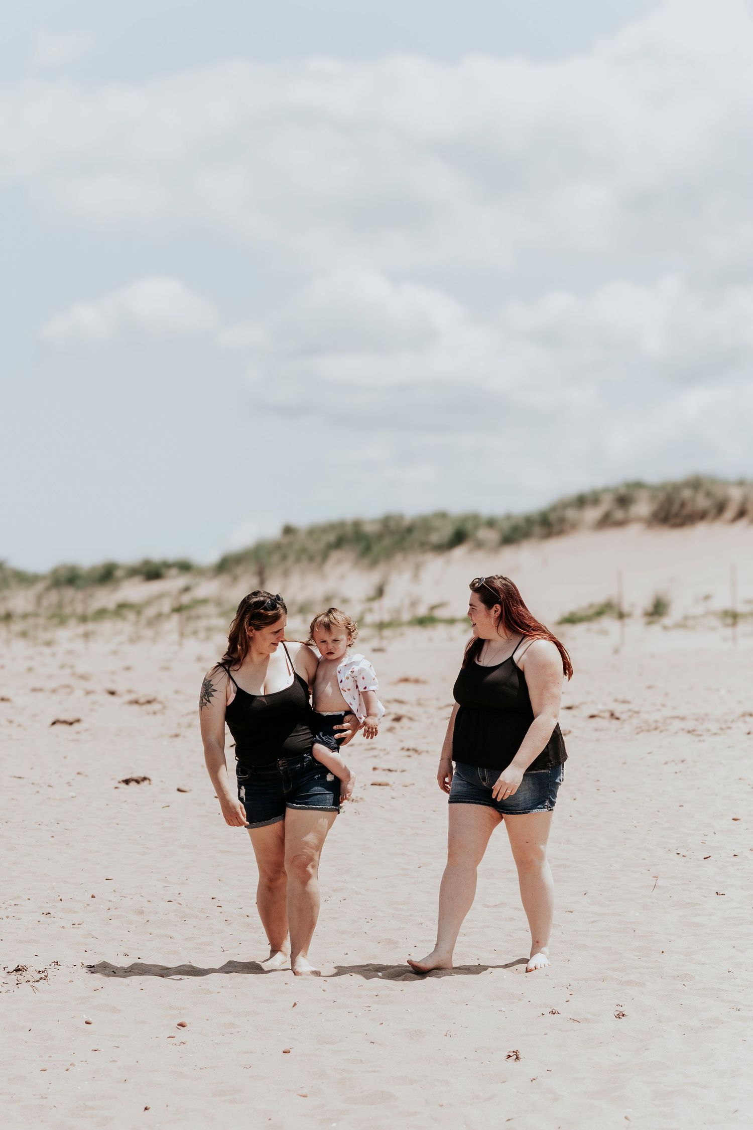 Two women in black tanks and denim shorts holding a toddler on a sunny beach.