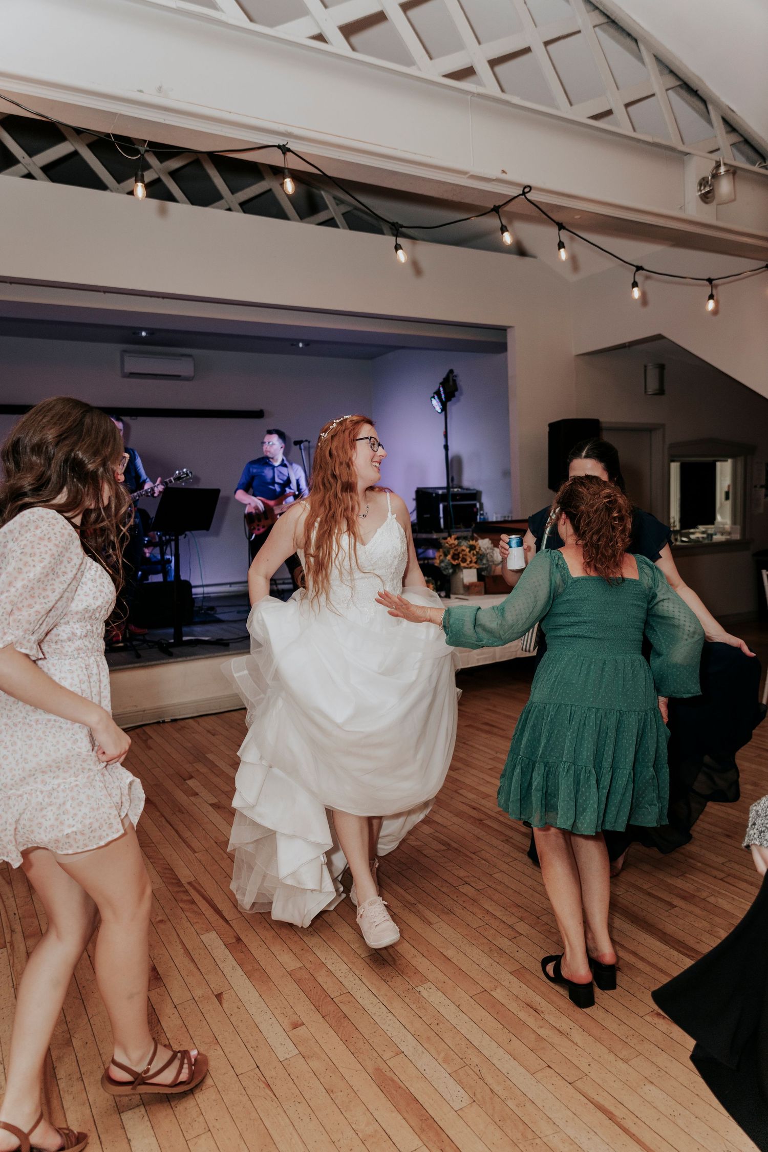 Bride in white gown dancing energetically with guests on wooden dance floor at wedding reception.