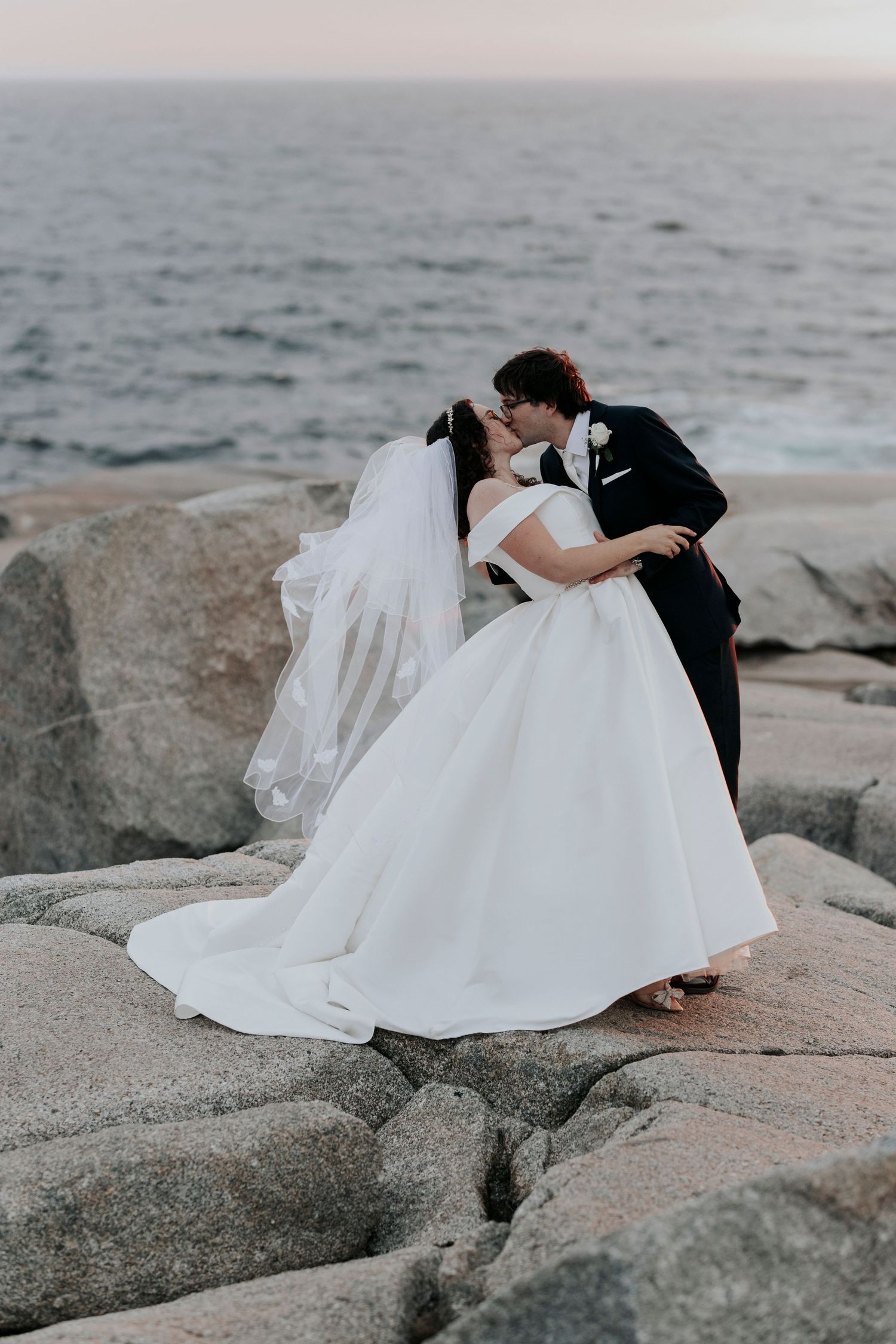 A romantic wedding moment captured on coastal rocks at sunset by the ocean.