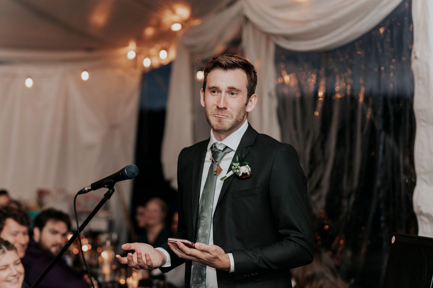Speaker gives wedding toast while standing at microphone under draped tent ceiling with string lights.