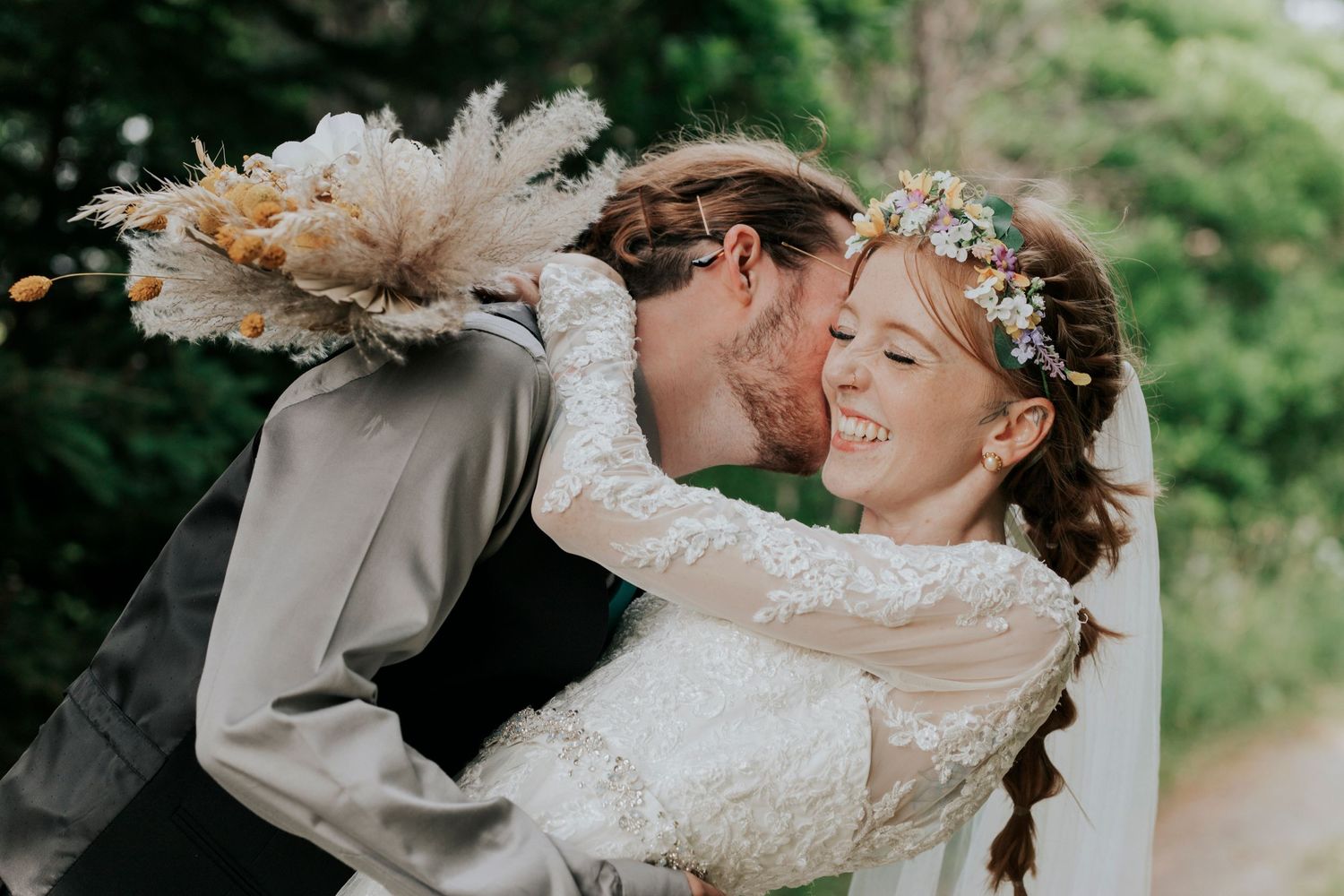 Joyful romantic moment captured outdoors with bride in lace dress and floral crown under natural greenery.