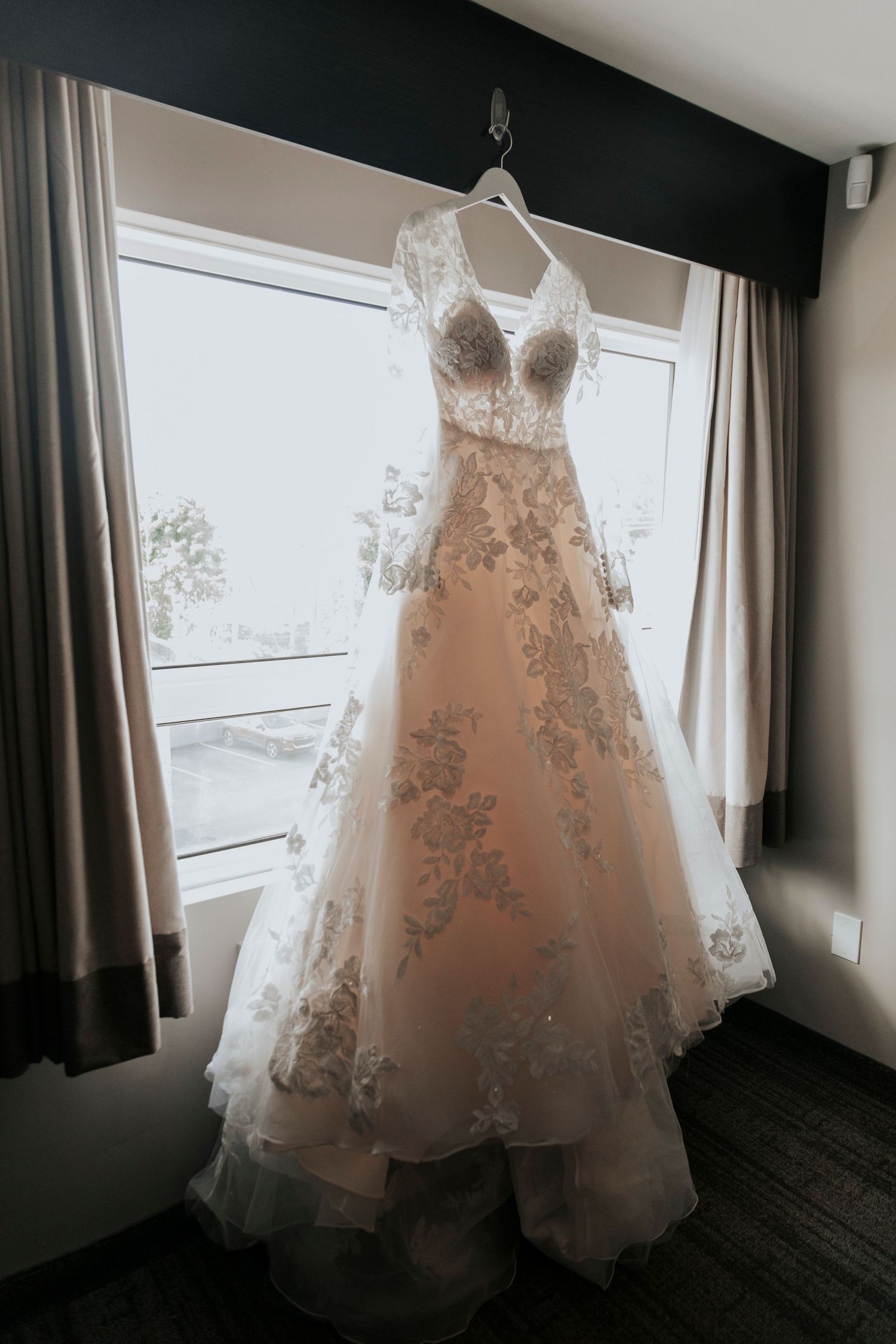 A delicate lace wedding dress with sweetheart neckline hangs by a window in natural light.