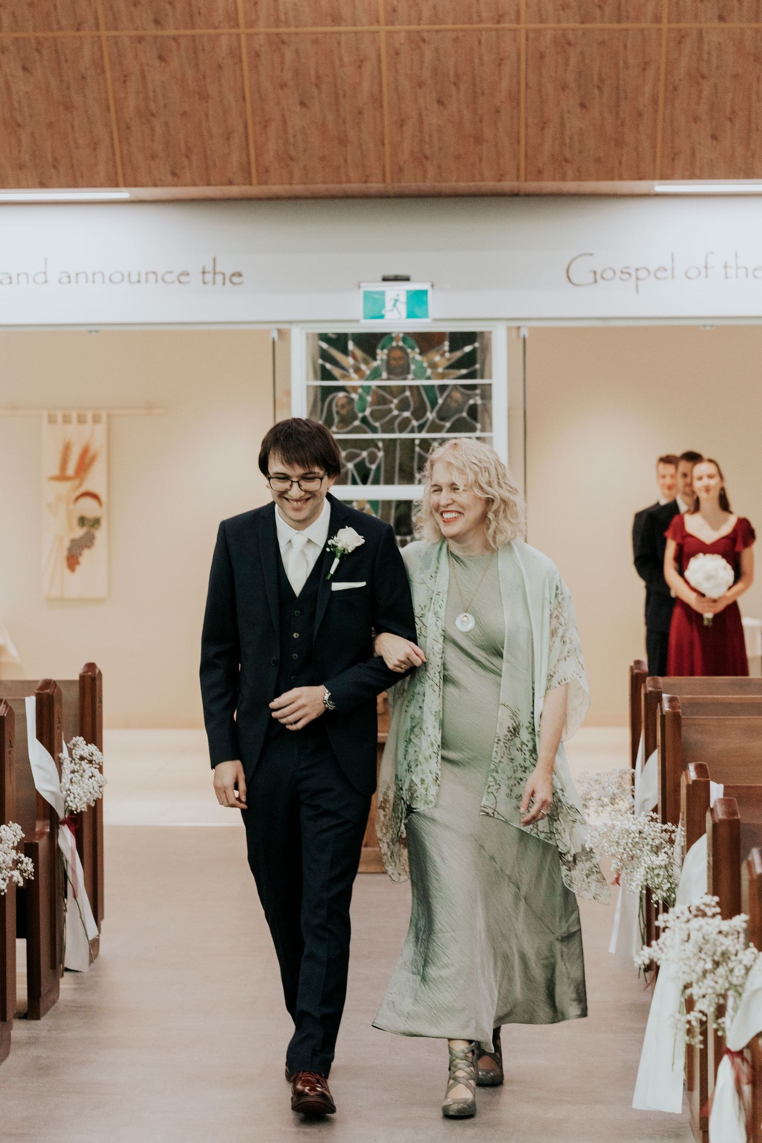 A young couple walks arm in arm down the aisle of a church decorated with white flowers and wooden pews.