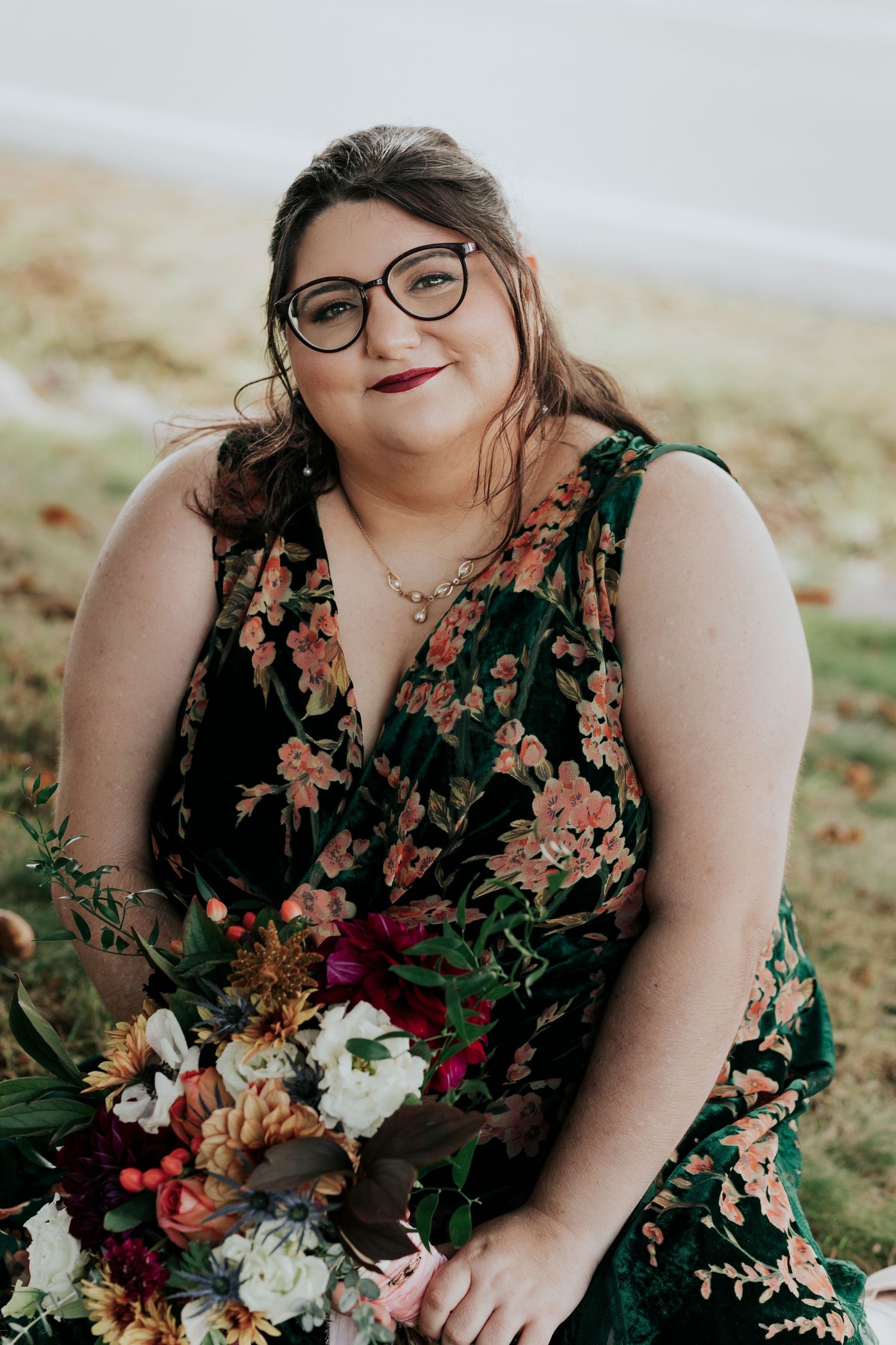 Bride in dark floral dress holding colorful bouquet seated outdoors near waterfront.