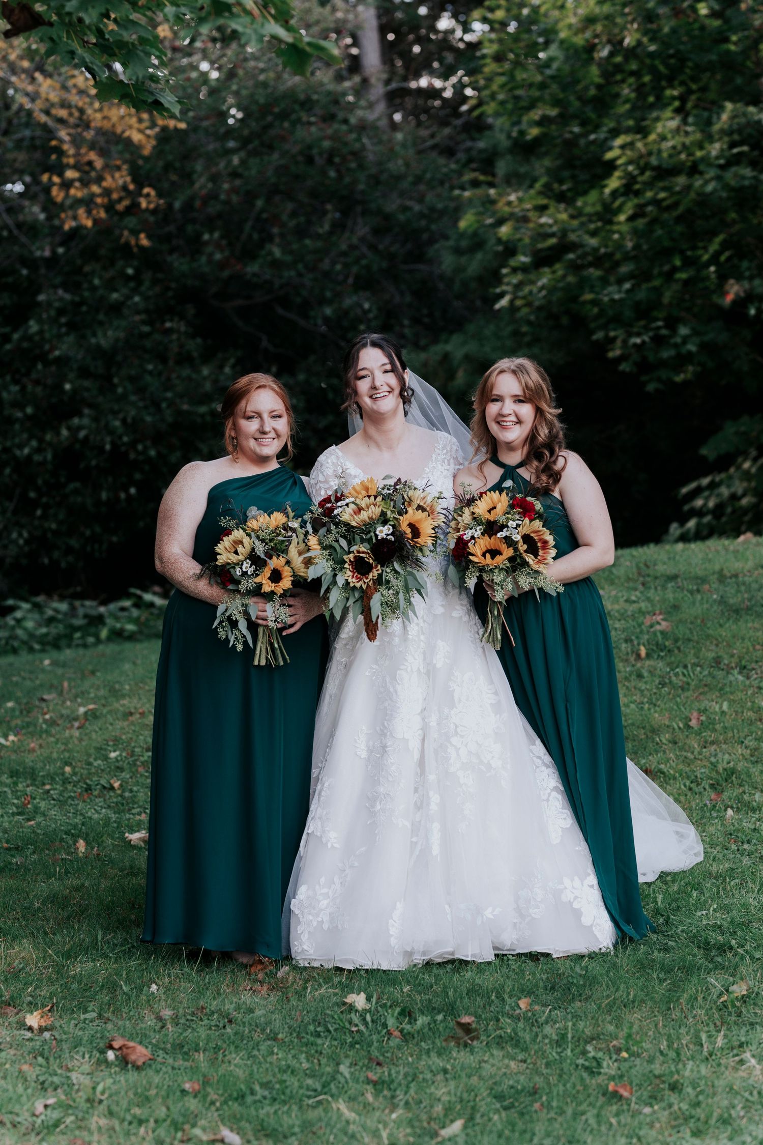 Bride poses with two bridesmaids in hunter green gowns holding sunflower bouquets outdoors.