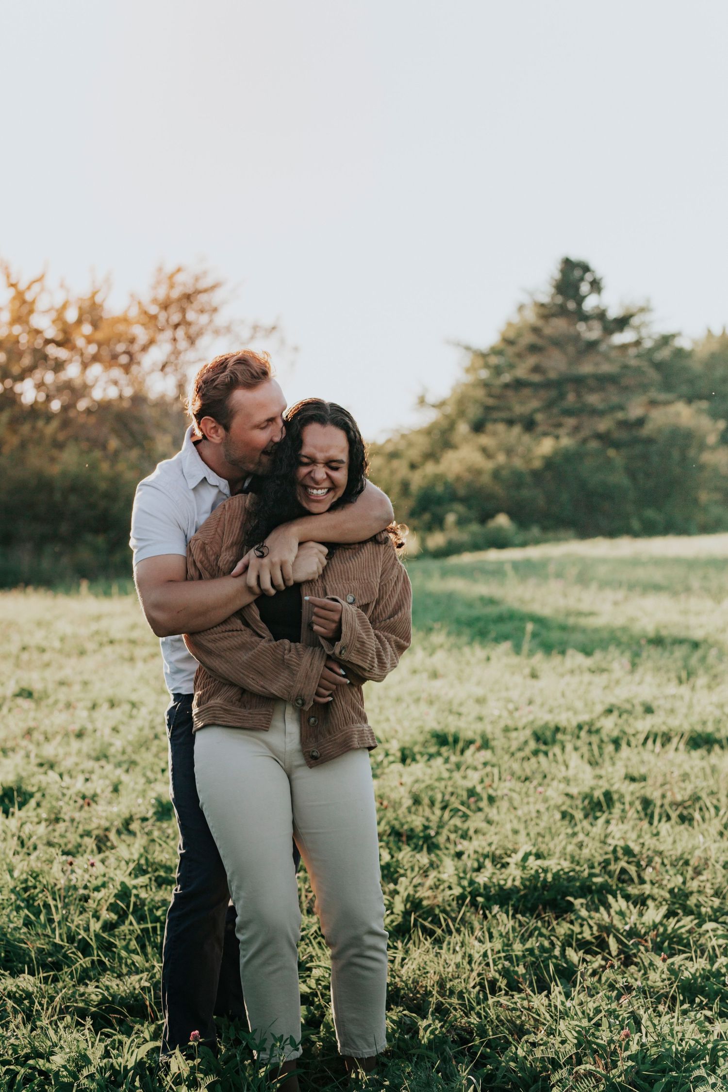 Man hugging woman from behind as she laughs joyfully in a sunlit green meadow at sunset.