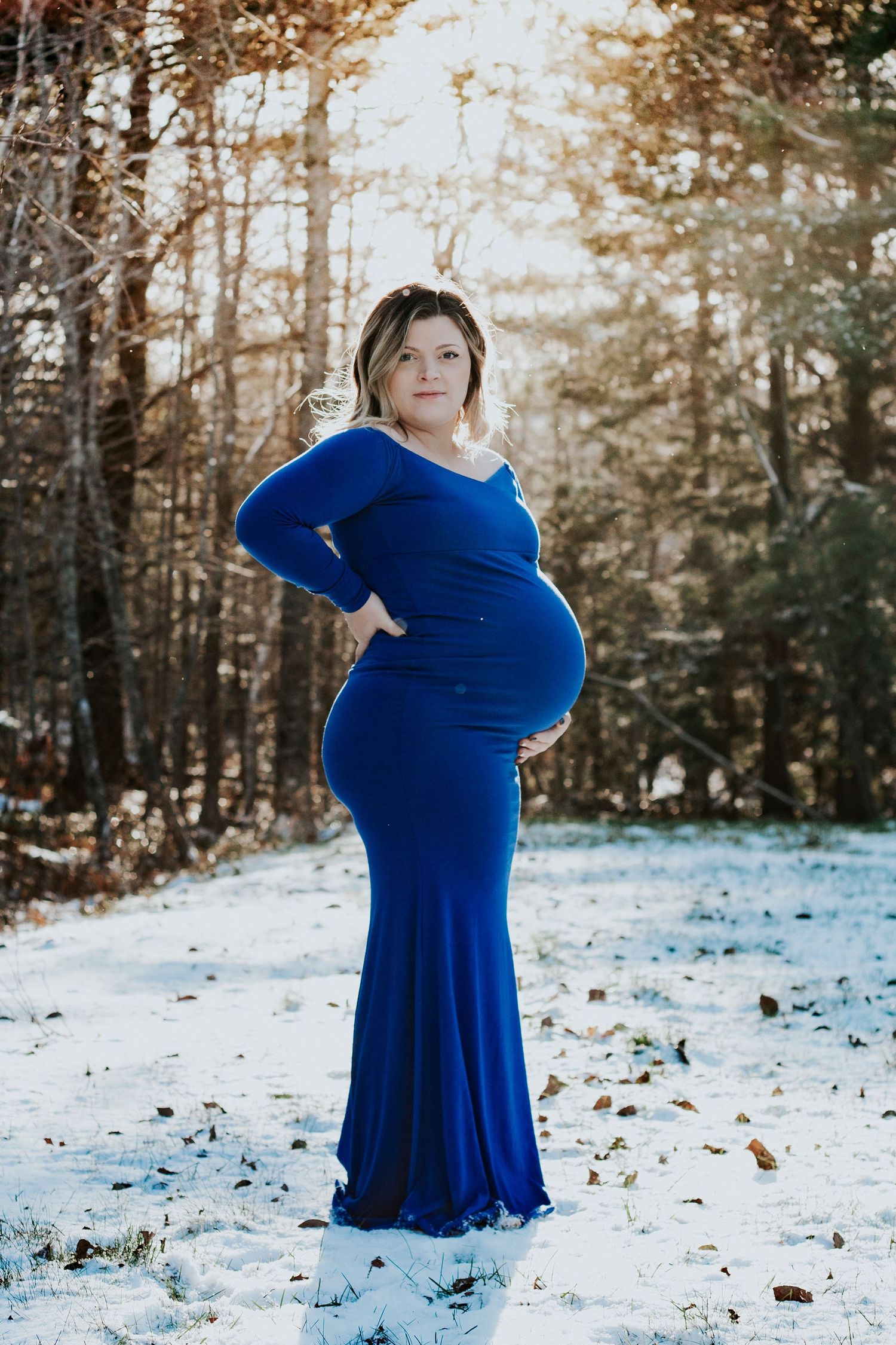 Pregnant woman in a royal blue gown poses in a snowy winter forest during a maternity photo session.