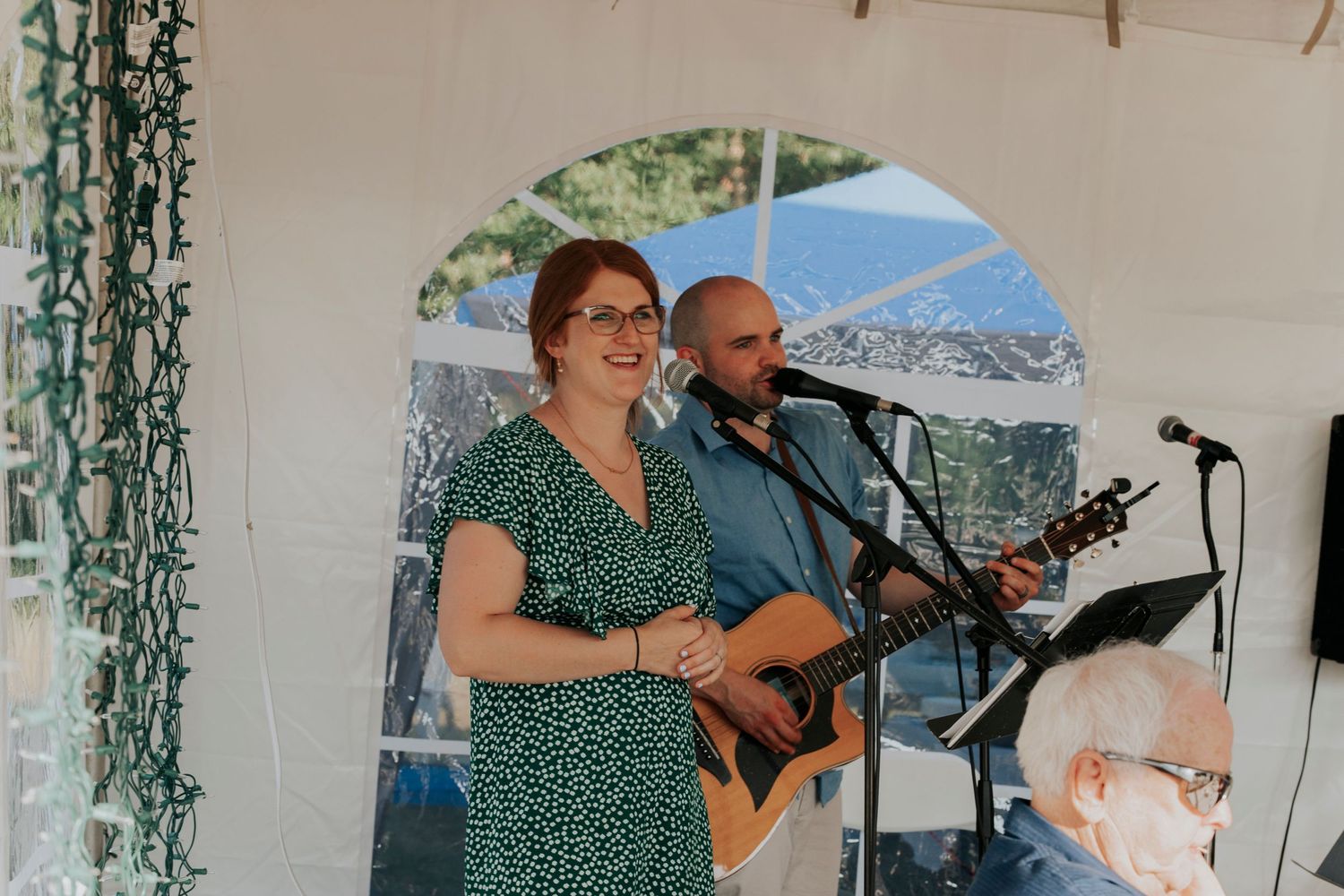 Musicians perform together under a white gazebo at an outdoor event with acoustic guitar and microphones.