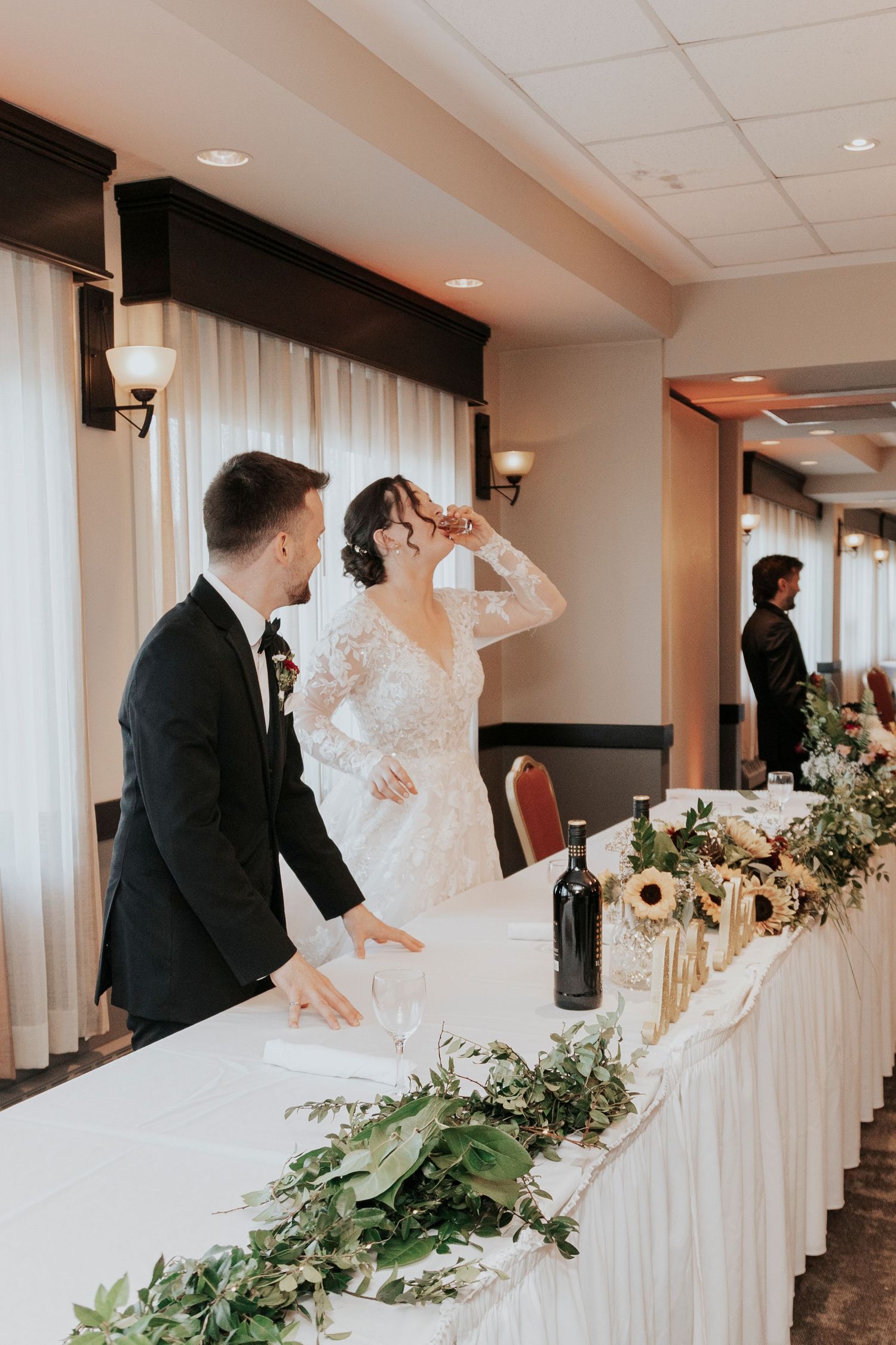 Couple stands at their reception head table adorned with greenery and floral decorations.