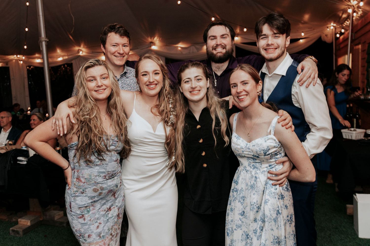 Wedding party group poses together under tent with string lights at evening reception.