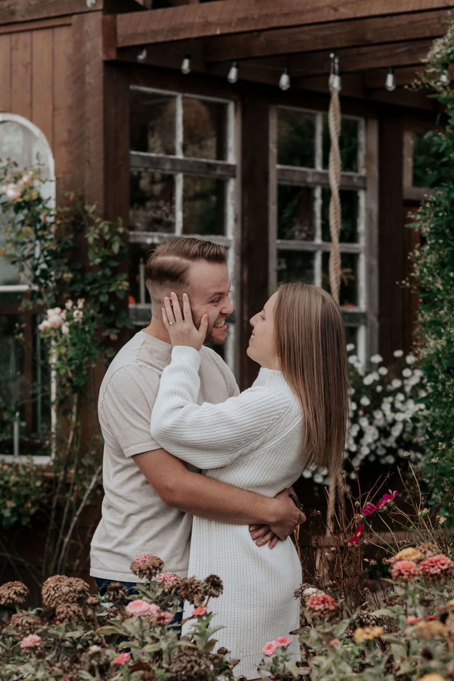 Couple embracing and smiling at each other in a romantic garden with flowers and rustic wooden building backdrop.