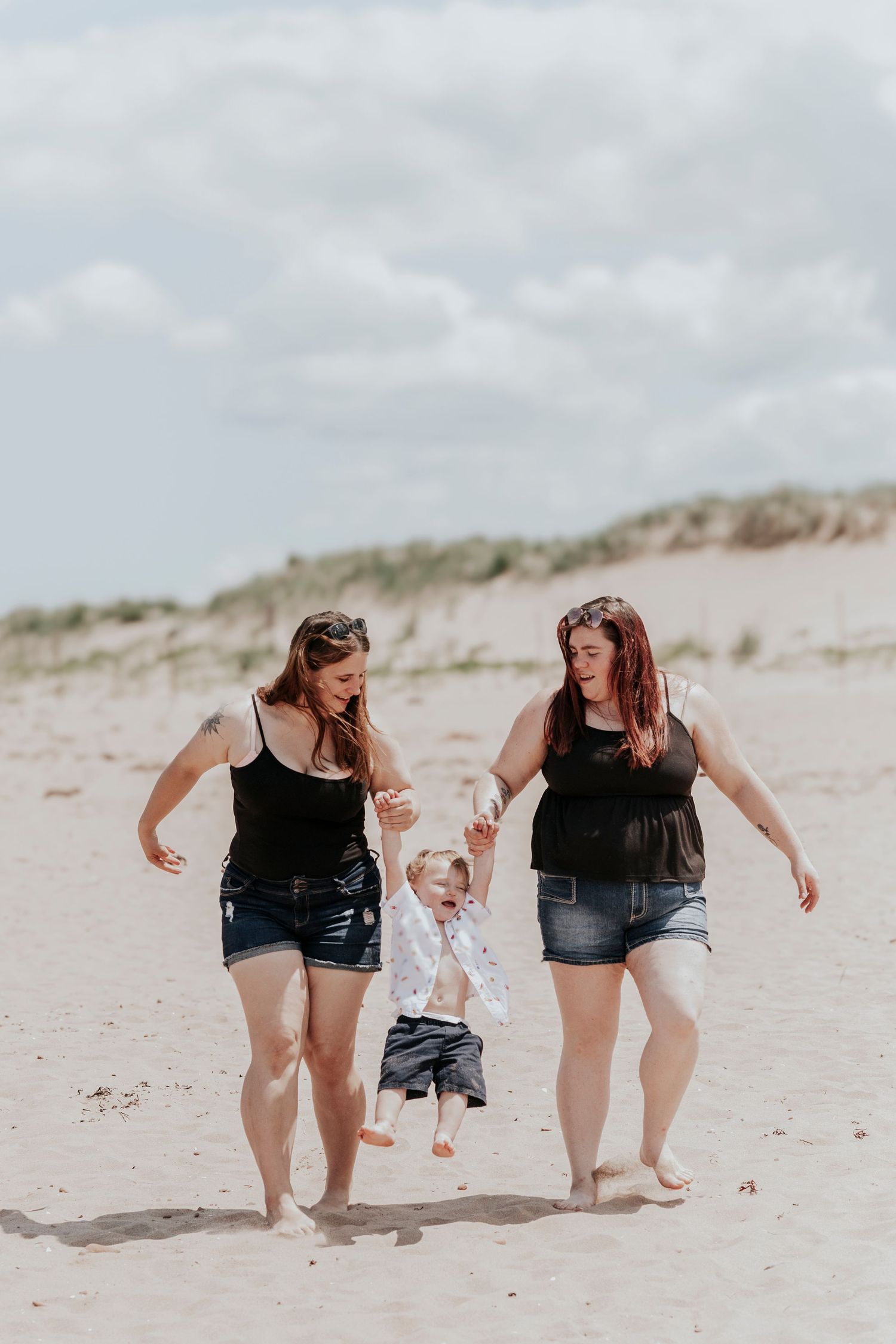 Two women lifting happy toddler by hands while walking on sandy beach together.