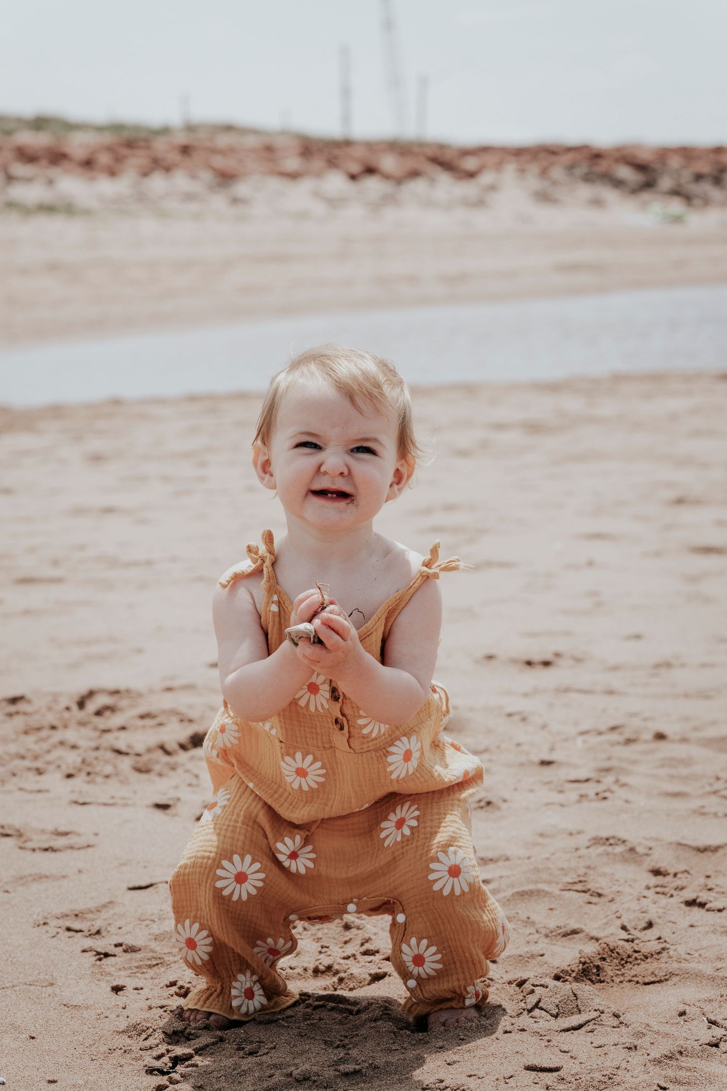 Smiling baby girl in daisy romper playing on sandy beach.