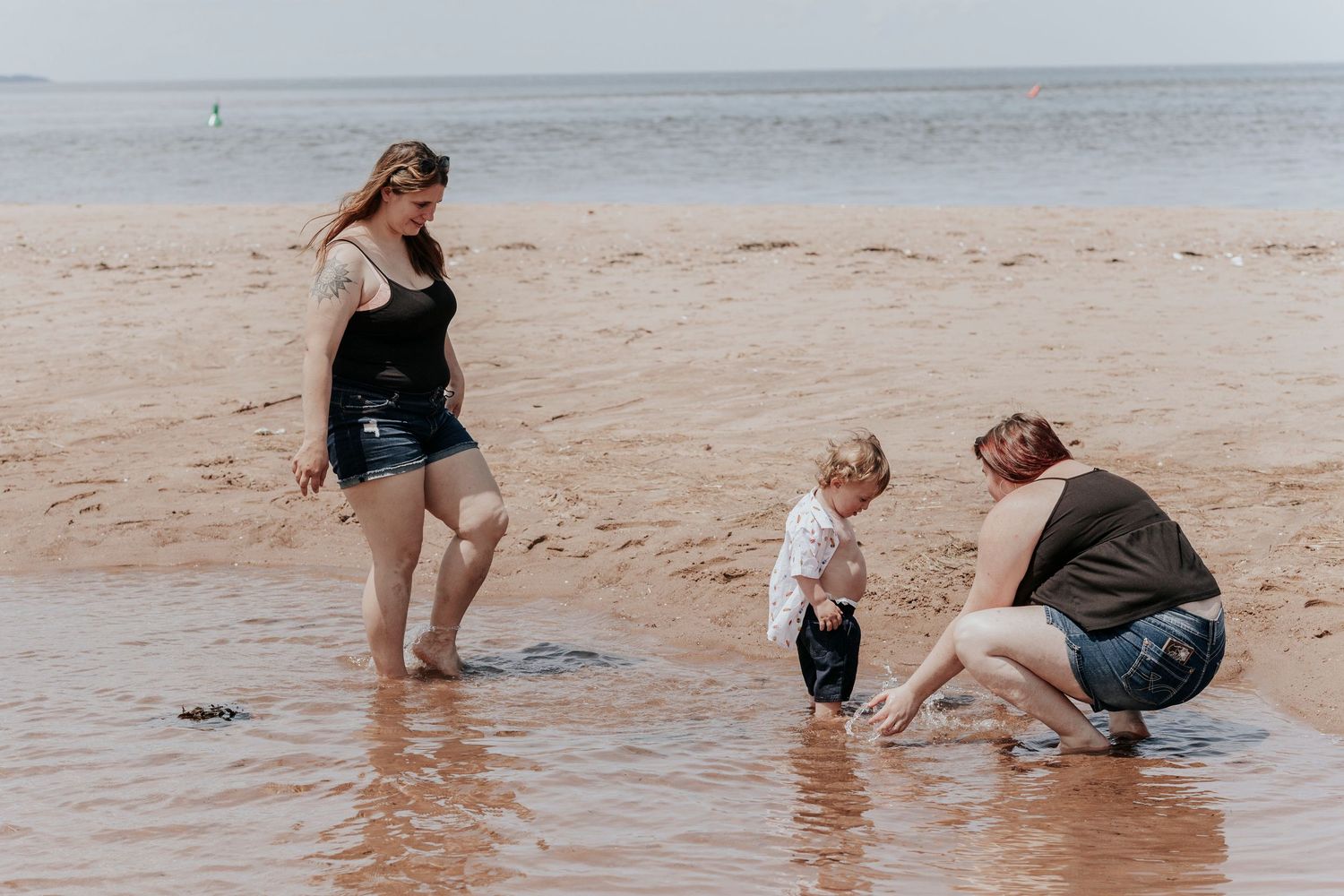 Family of three with toddler playing together in shallow water on a red sand beach.
