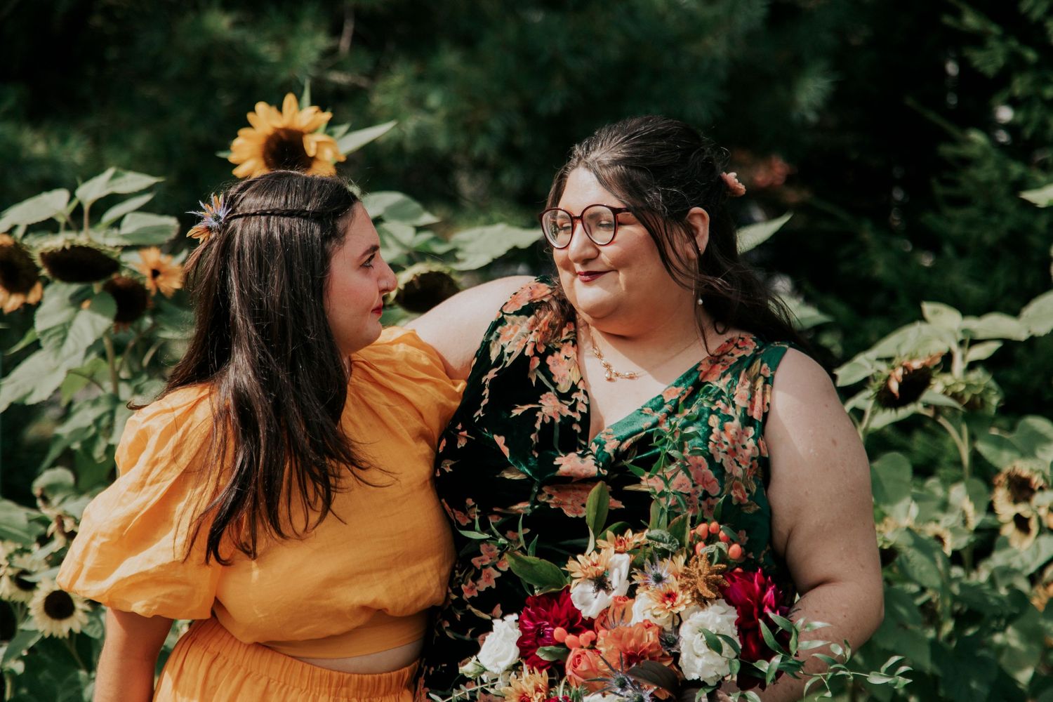 Two sisters share a joyful moment in a garden with sunflowers and colorful florals, one in yellow and one in floral print.