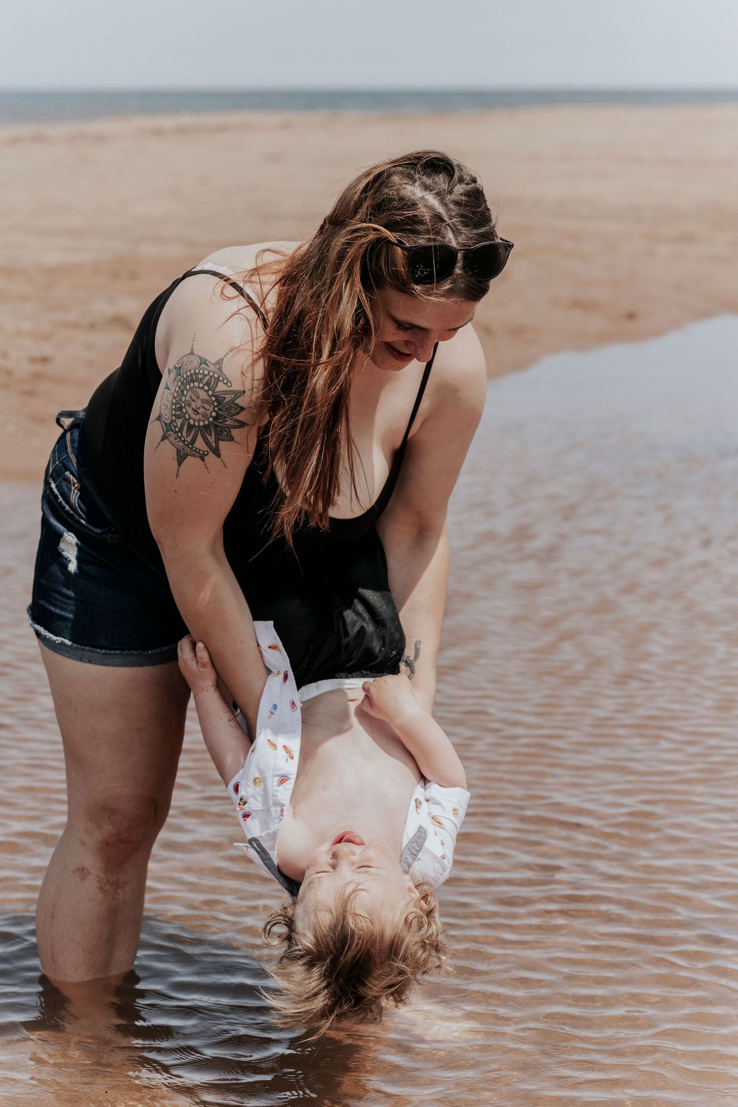 Tattooed mother playfully dipping laughing toddler upside down into shallow beach water.