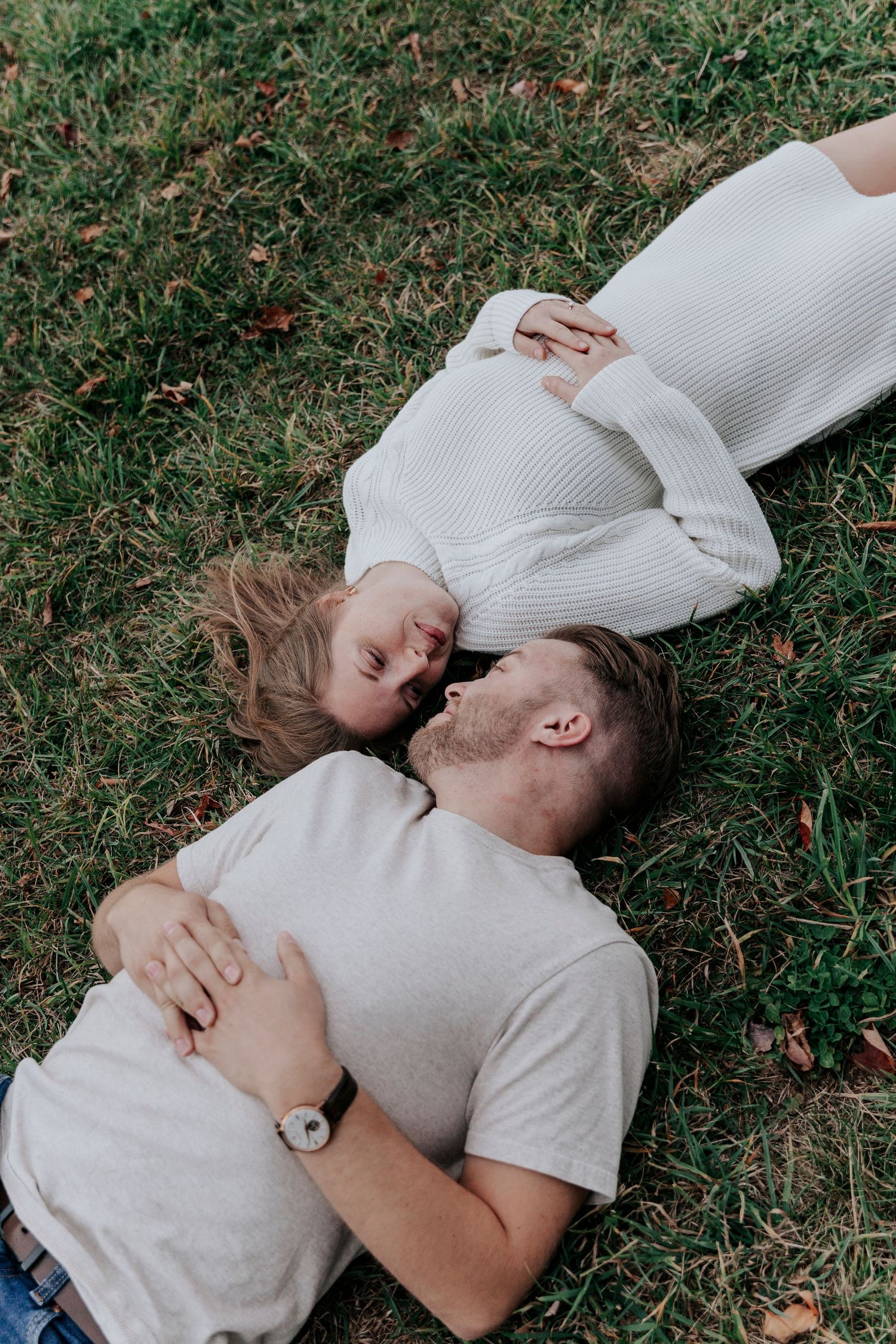 Couple lying on green grass laughing together in a relaxed outdoor engagement session.