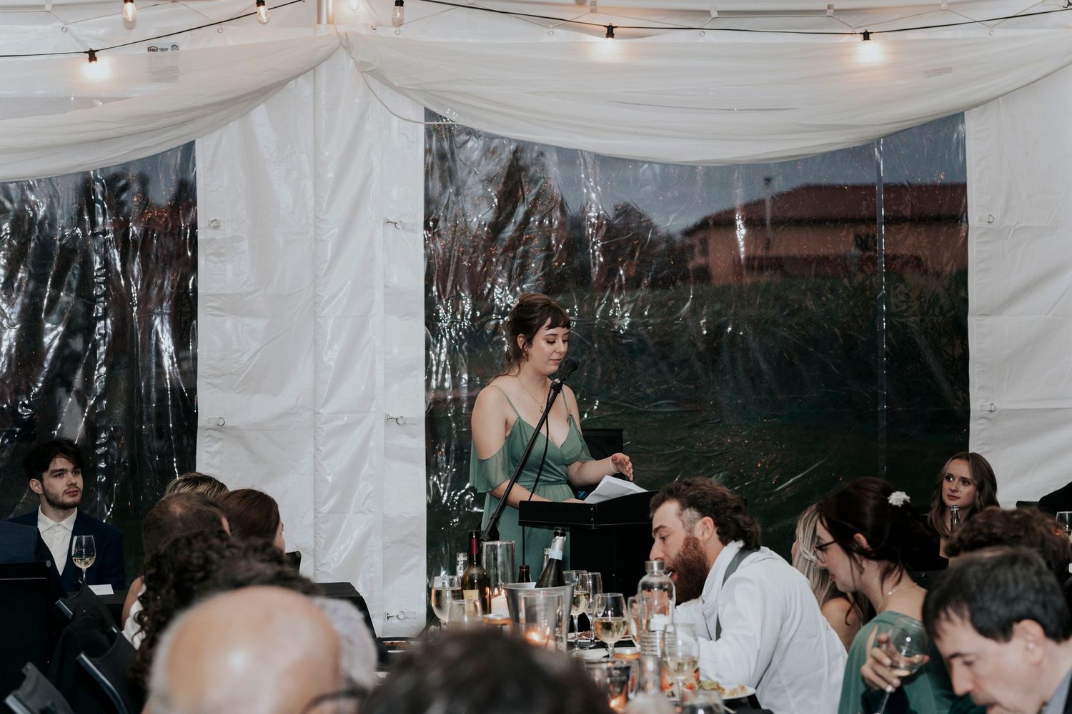 Wedding guests gathered in a tent-covered venue listen to speeches during reception celebration.
