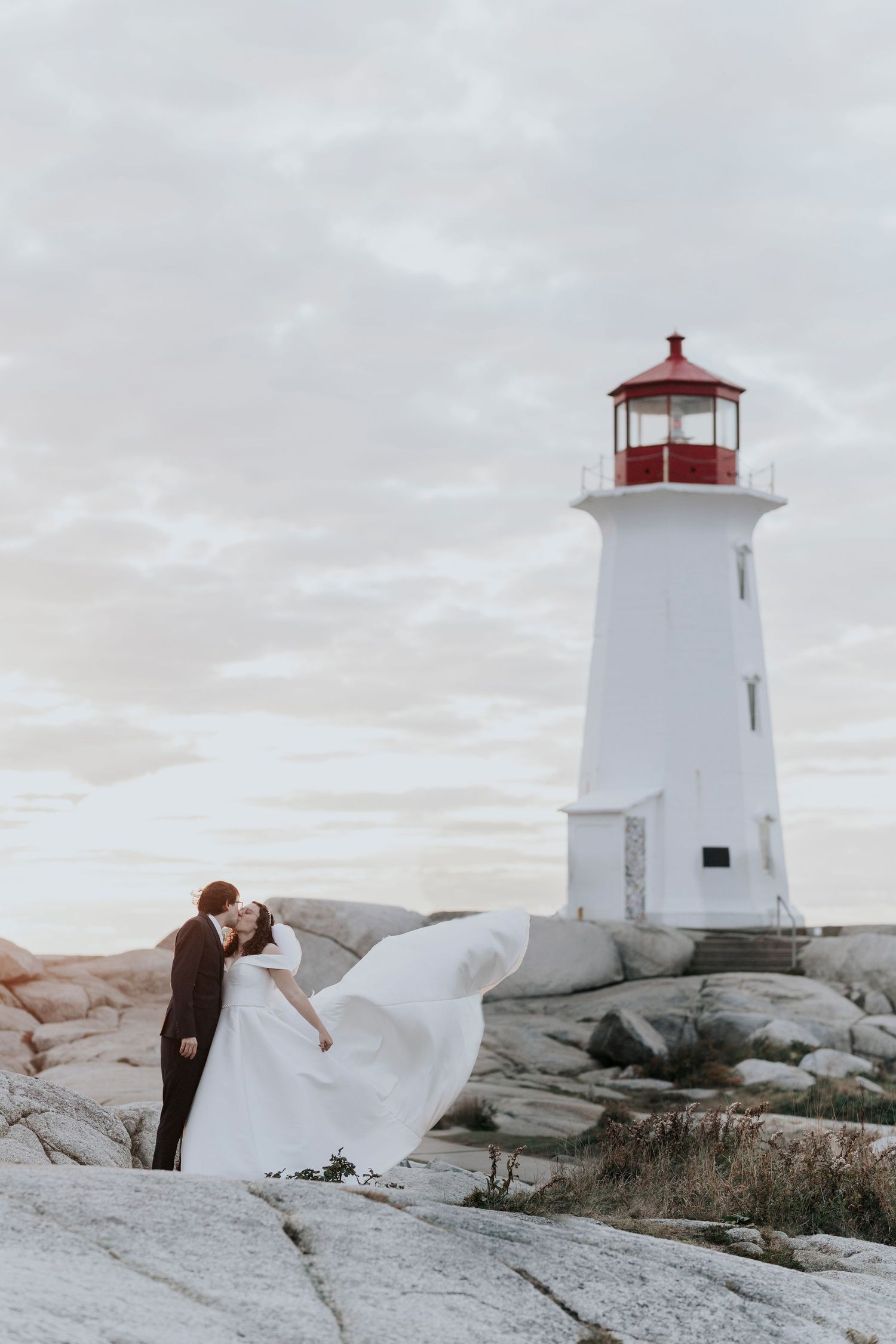A romantic wedding photo at Peggy's Cove lighthouse in Nova Scotia with bride's dress flowing in the wind against gray skies.