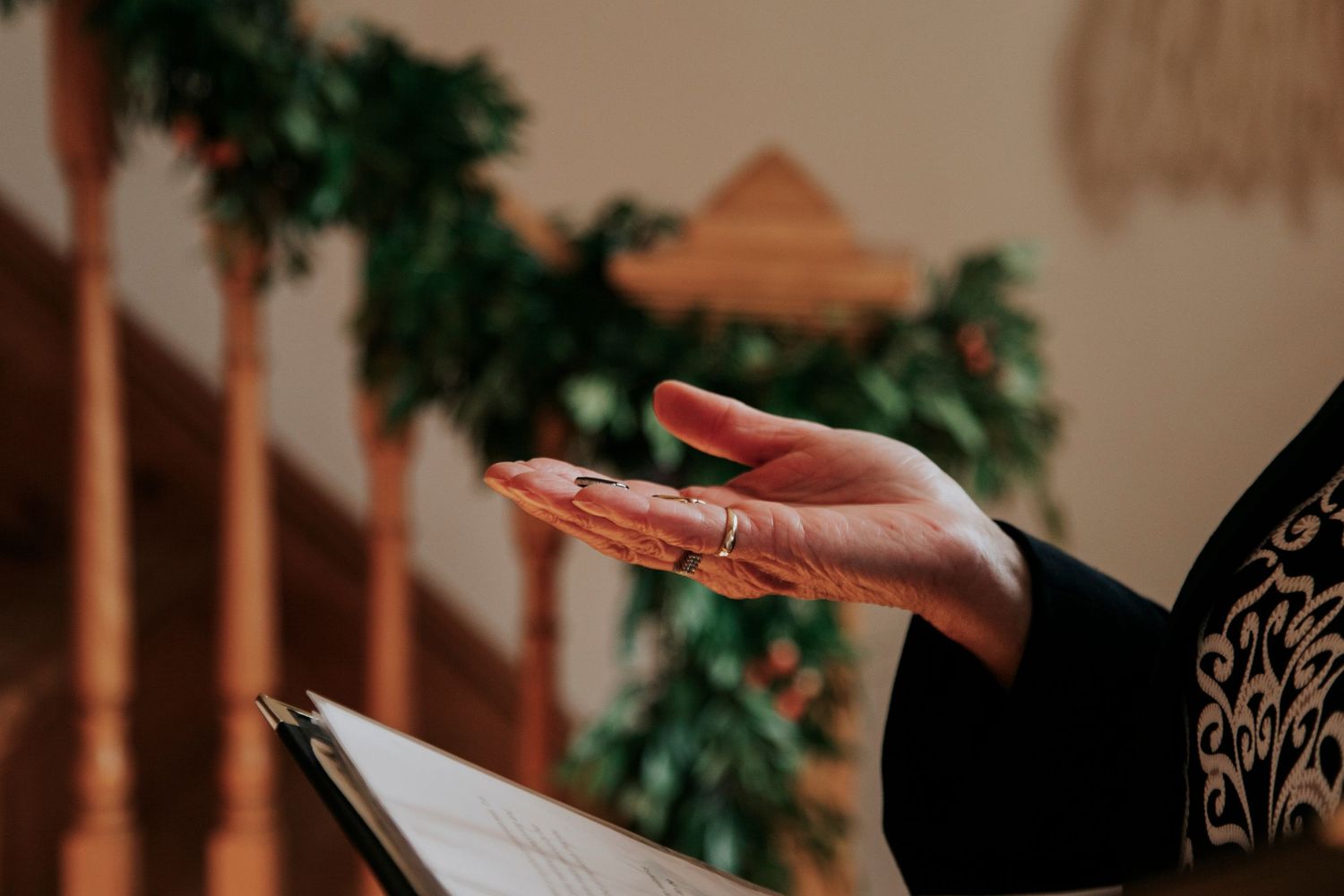 A raised hand extends upward in prayer while holding a religious book against a blurred background with greenery.