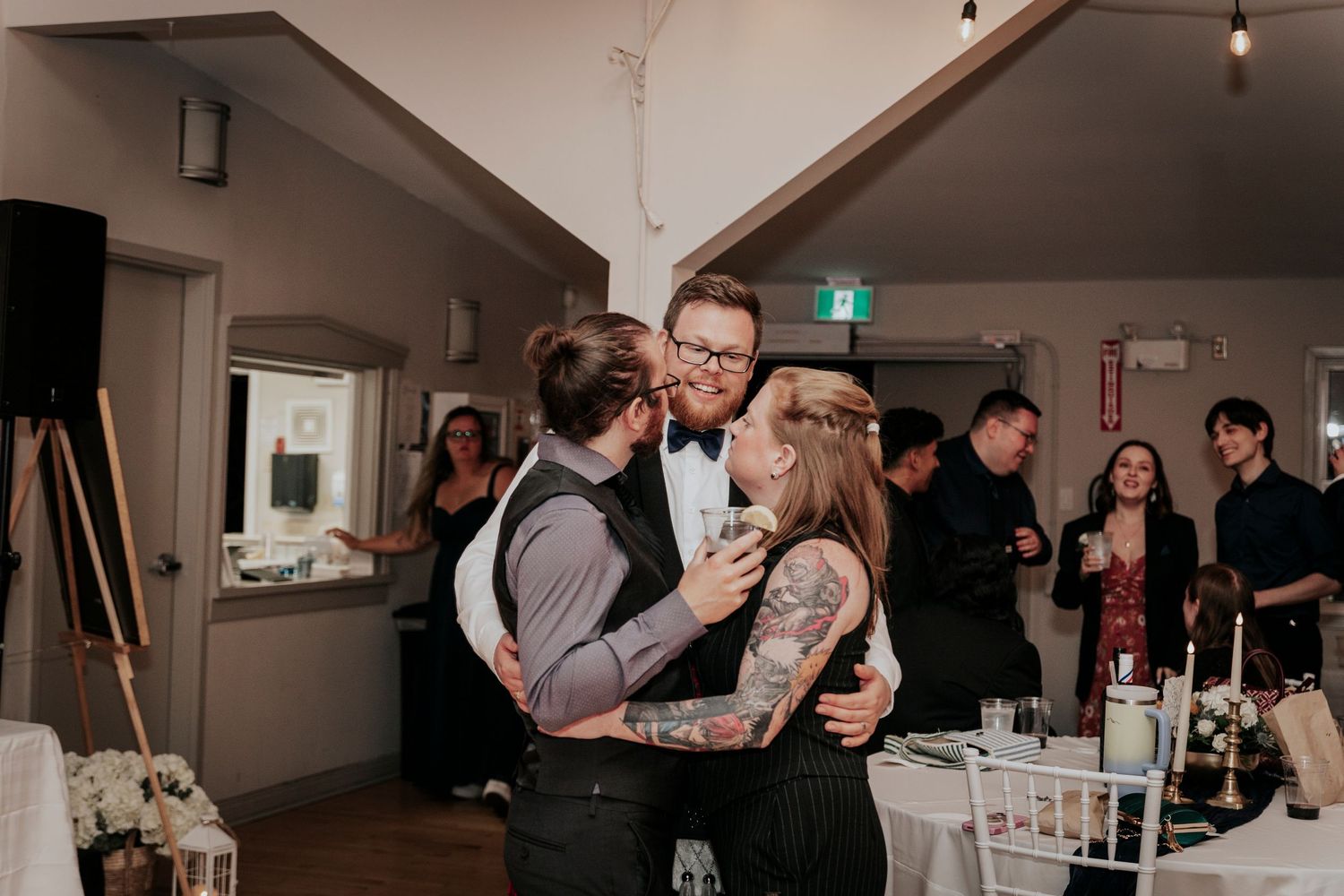 Groom in bow tie sharing a joyful group hug with two guests at wedding reception.