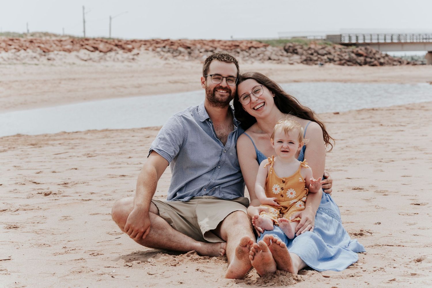 Happy family of three sitting together on sandy beach, parents smiling with baby girl in yellow floral romper.