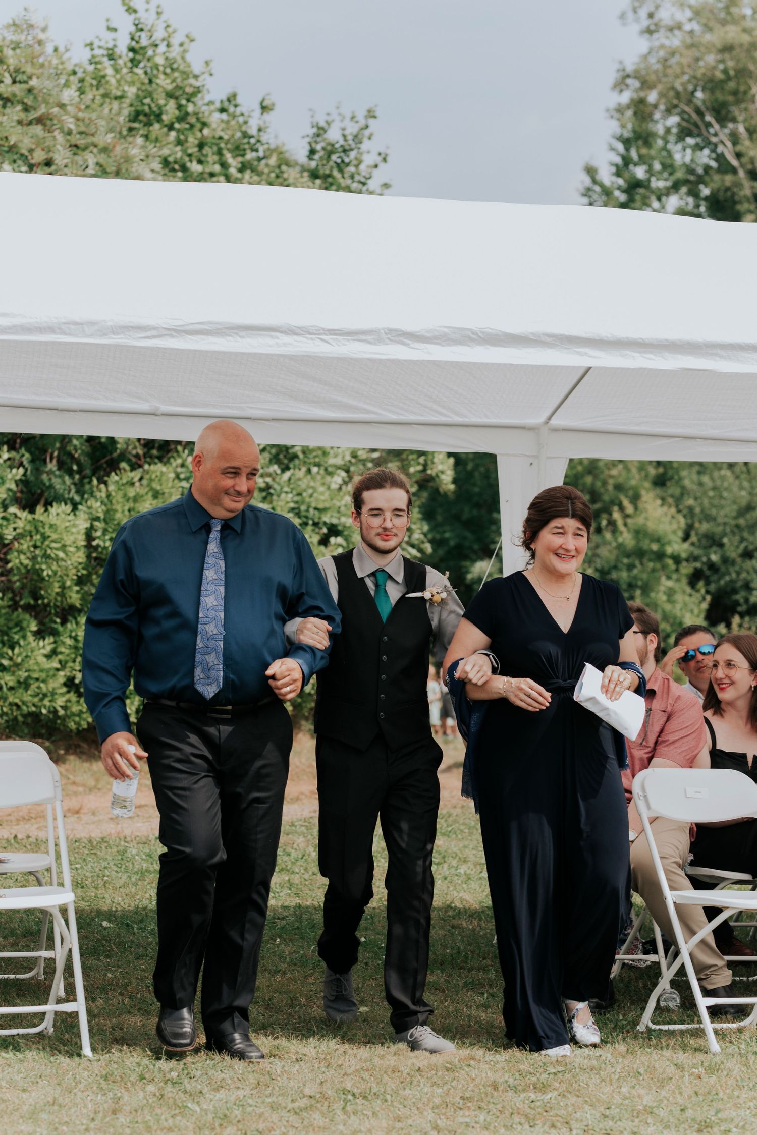 The groom and his parents walk down the aisle during an outdoor wedding ceremony under a white tent canopy on a grassy lawn.