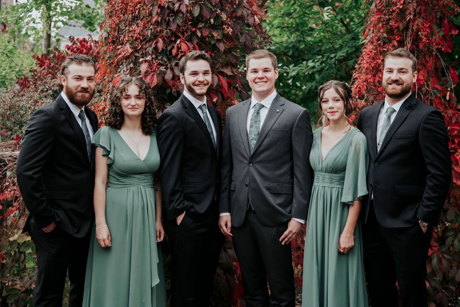 Wedding party in sage green dresses and dark suits posed against red foliage.
