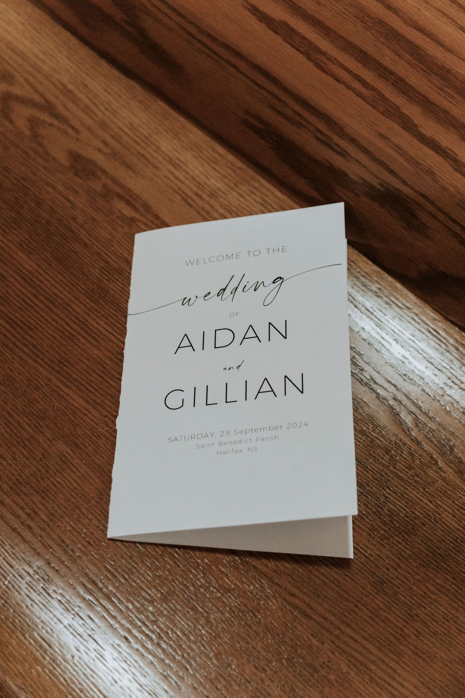 A minimalist white wedding program for Aidan and Gillian sits on a wooden surface.