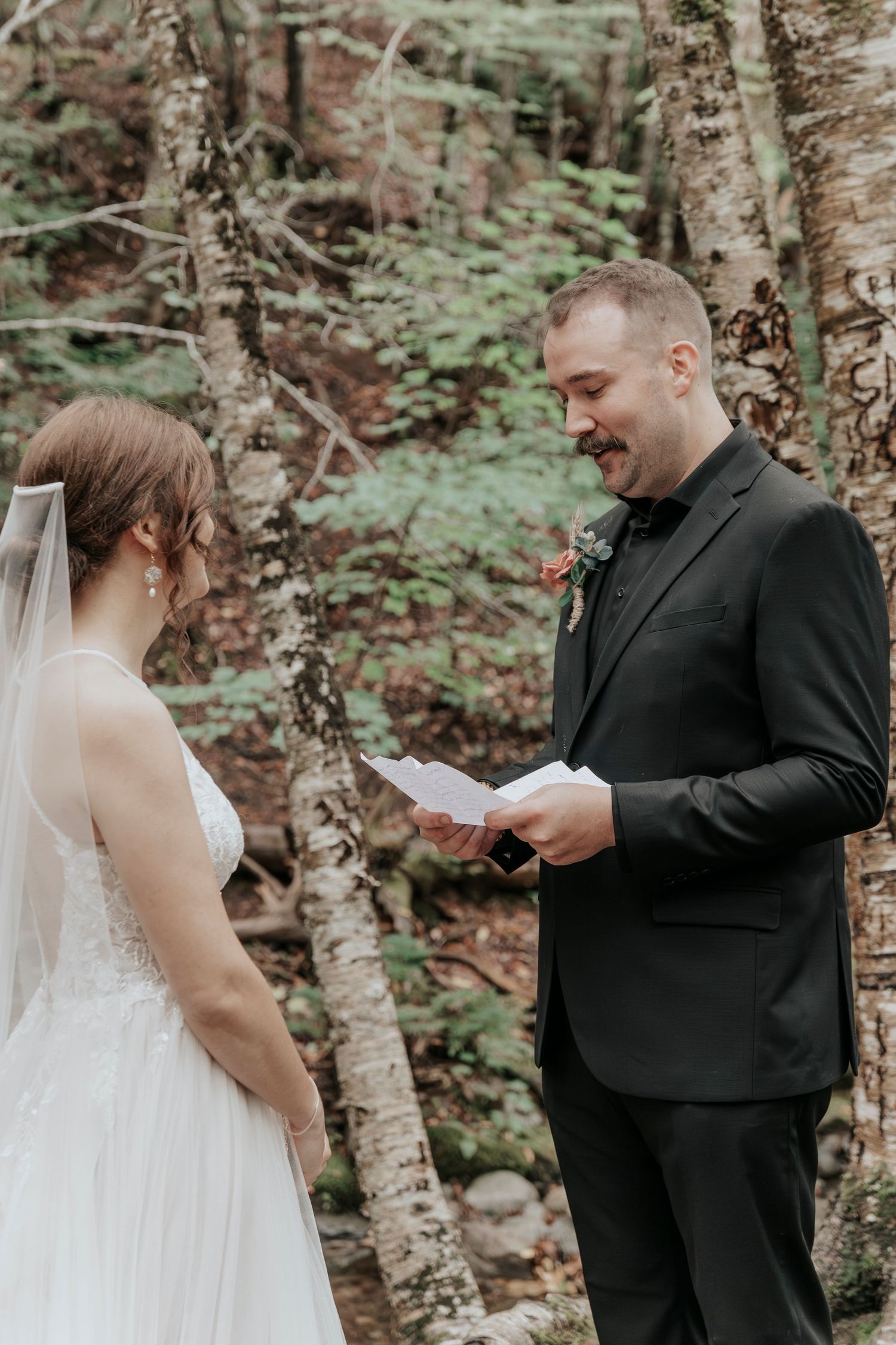 Groom reads heartfelt wedding vows from paper to his bride during an intimate forest elopement ceremony.