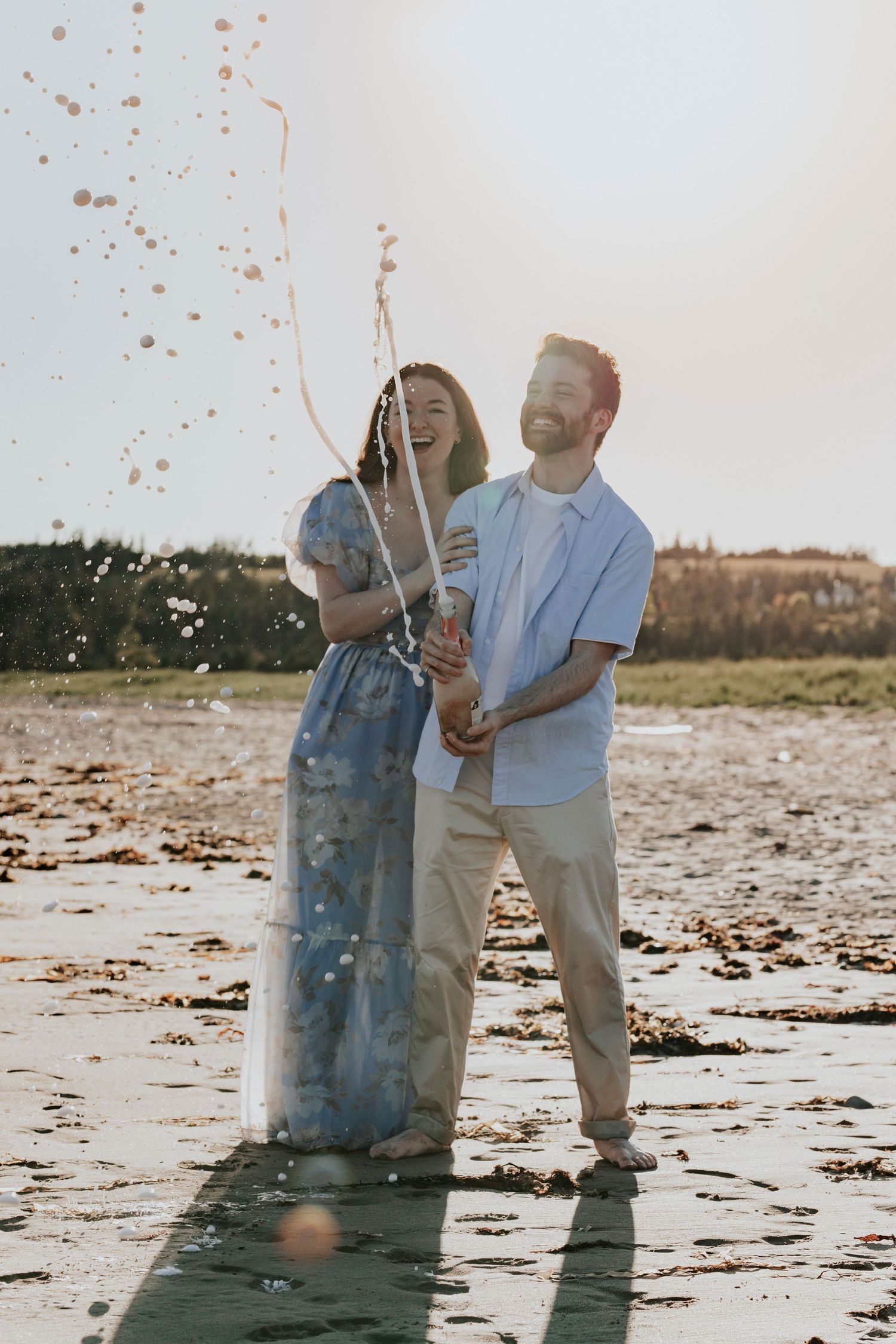 Joyful couple popping champagne bottle on a beach, celebrating with confetti and bubbles flying.